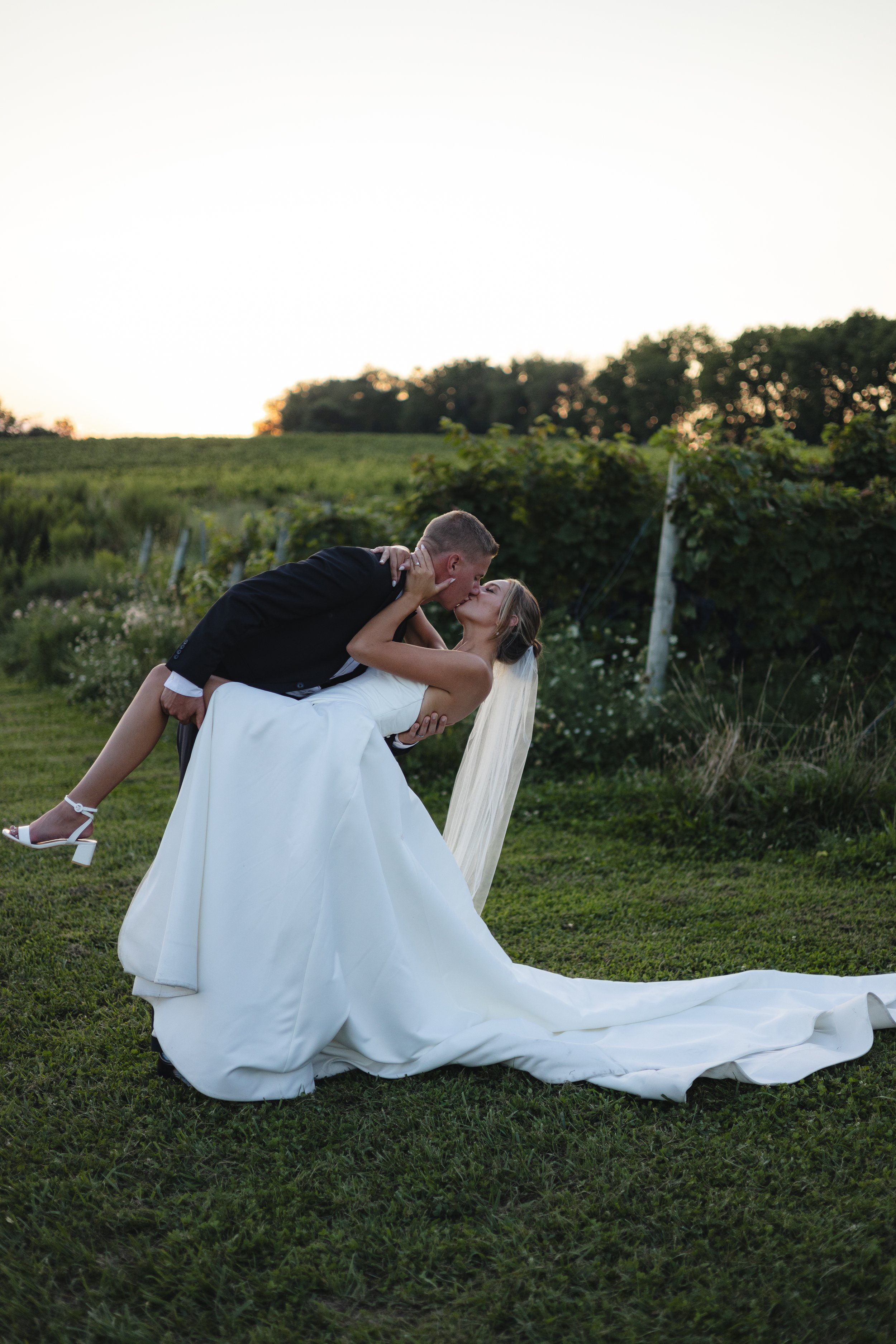 A bride and groom sharing a kiss outdoors during sunset, with the groom holding the bride in a dip. The bride is wearing a white wedding gown and veil, and the groom is in a suit. The background features green fields, trees, and a clear sky.