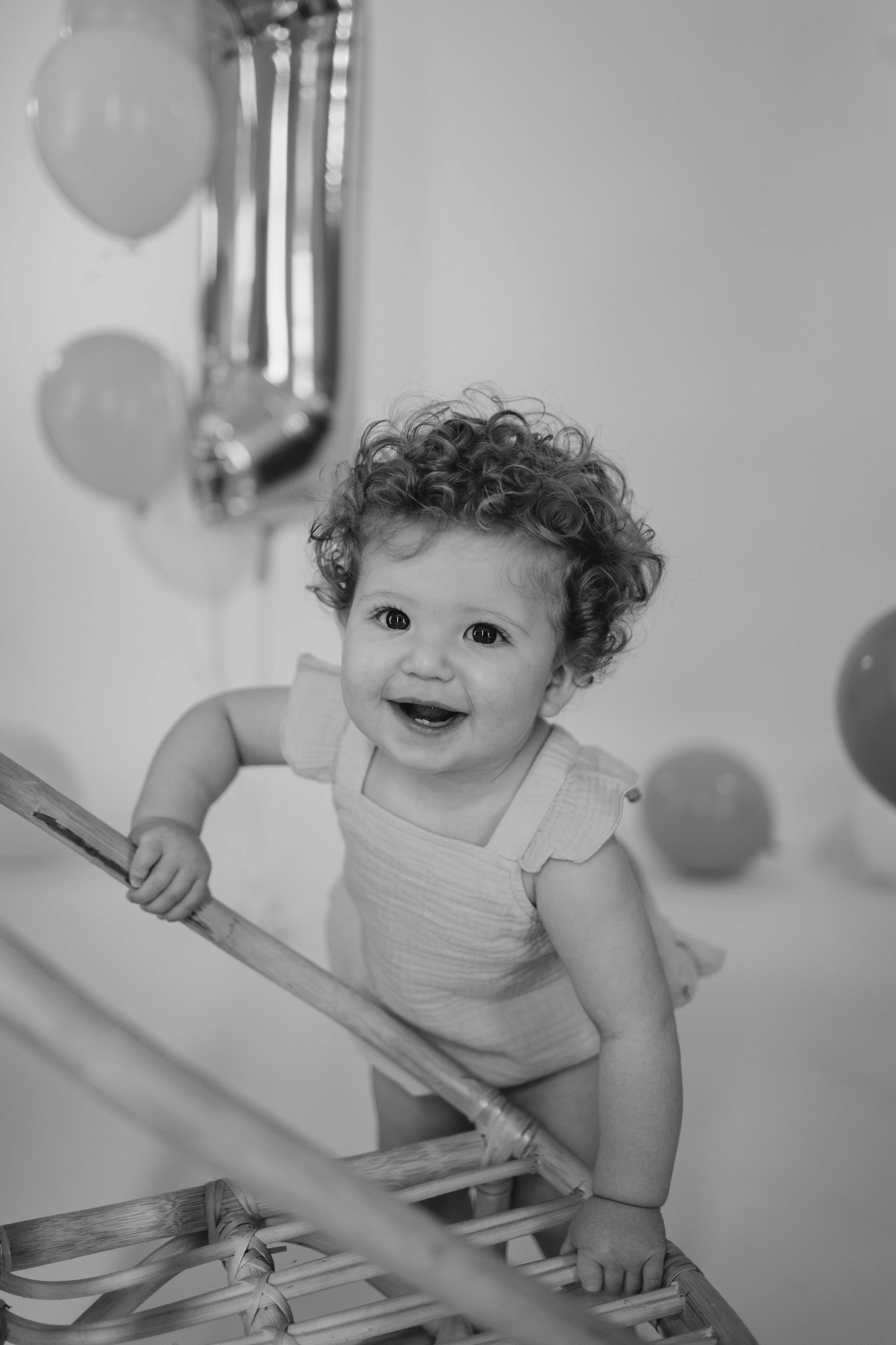 Black and white photo of a young child with curly hair smiling and holding onto a wooden railing, with an out-of-focus balloon and wall in the background.