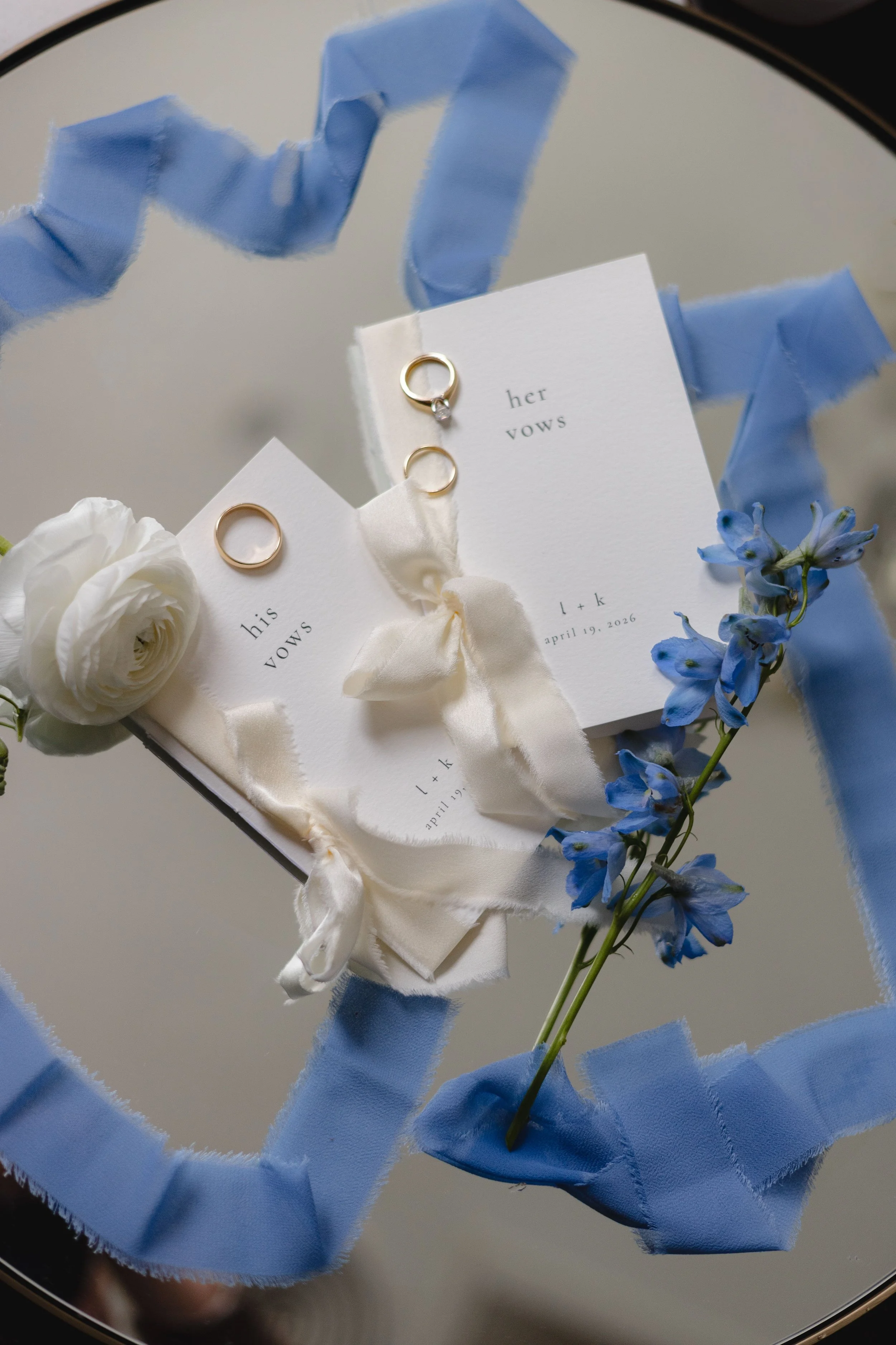 Wedding rings on white cards with vows, surrounded by white flowers and blue ribbon, on a reflective surface.