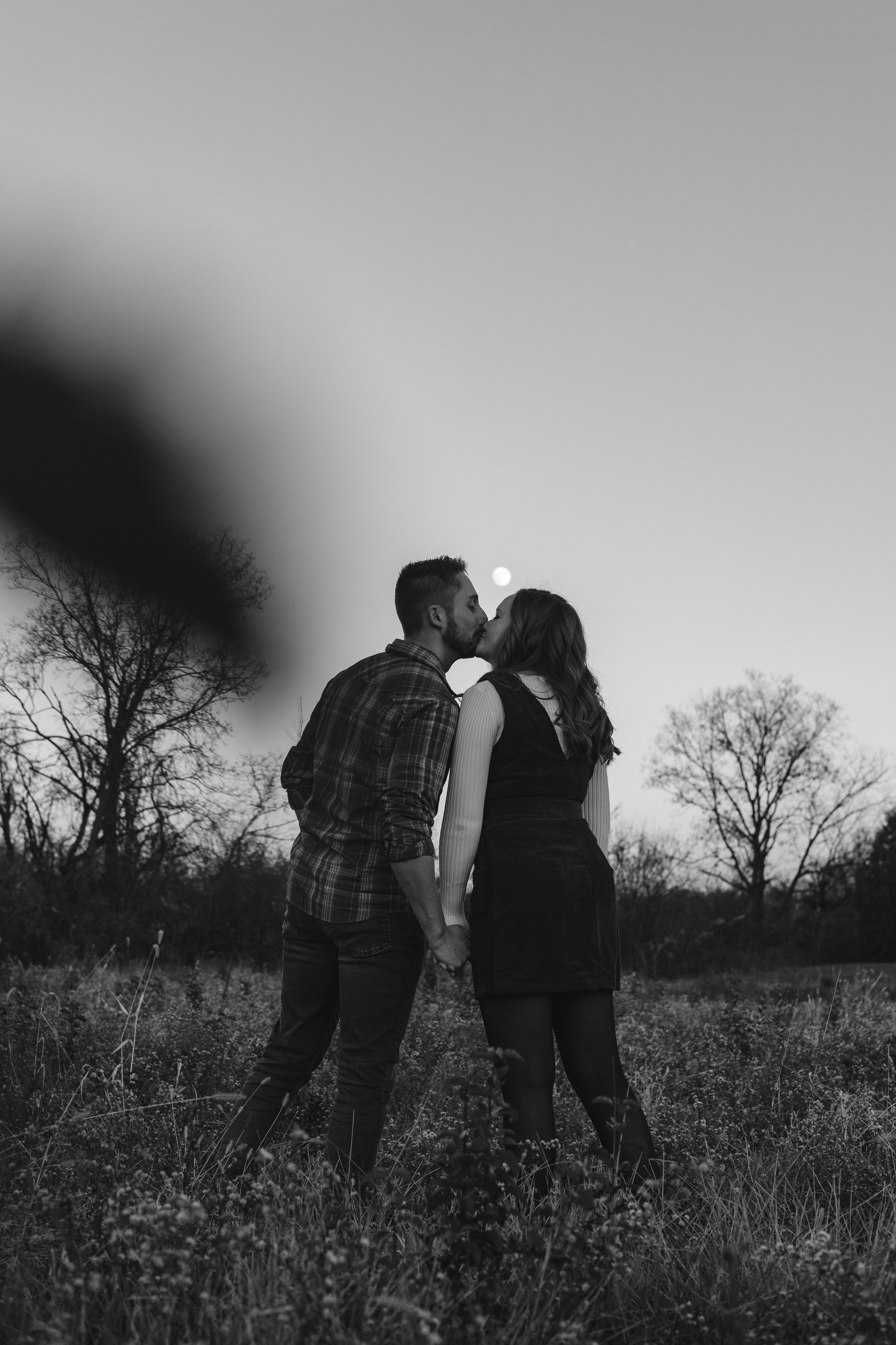 A black and white photograph of a couple sharing a kiss outdoors during sunset, with the moon visible in the sky and trees in the background.