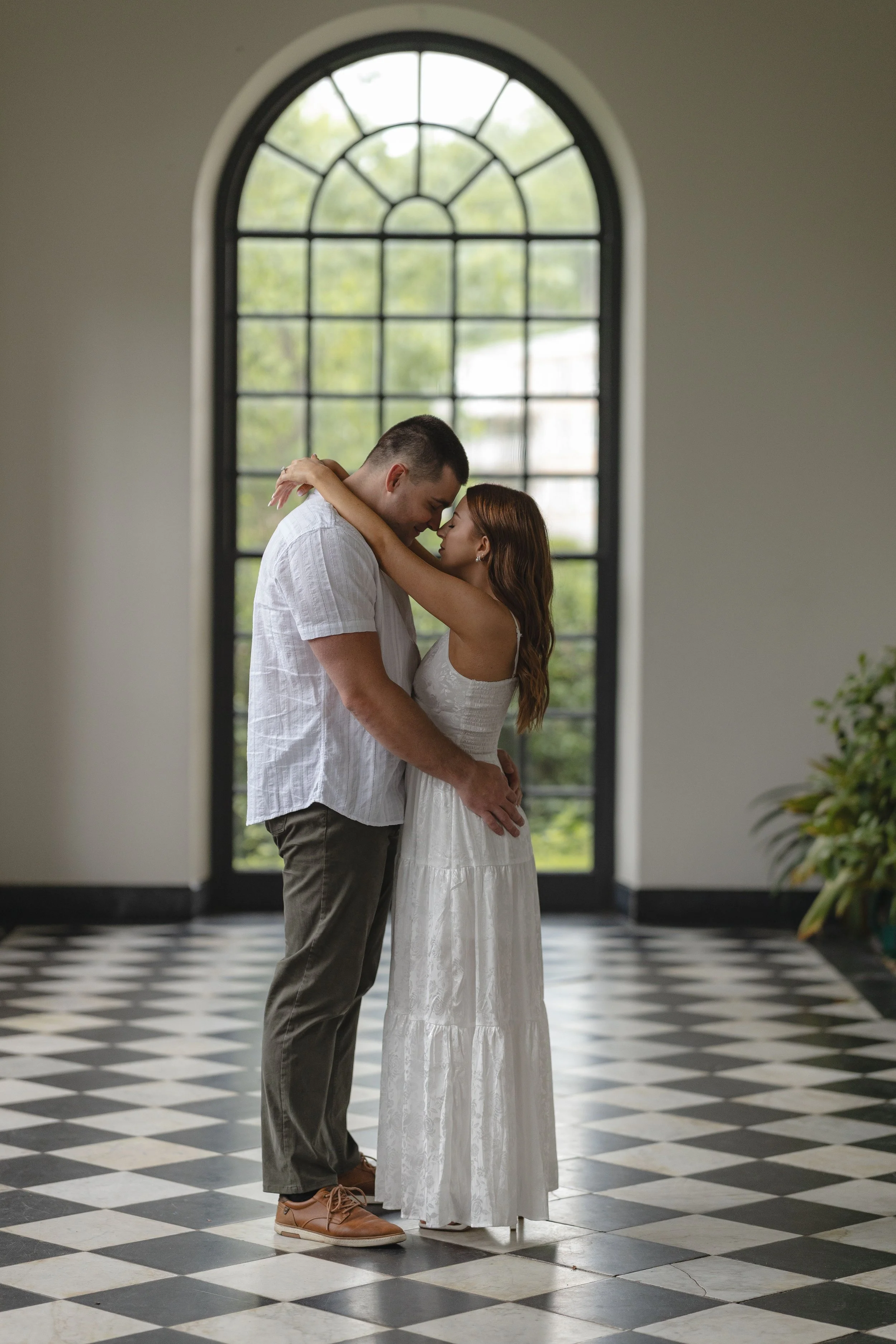 A couple embraces in front of a large arched window, with a black and white checkered floor and green trees outside.