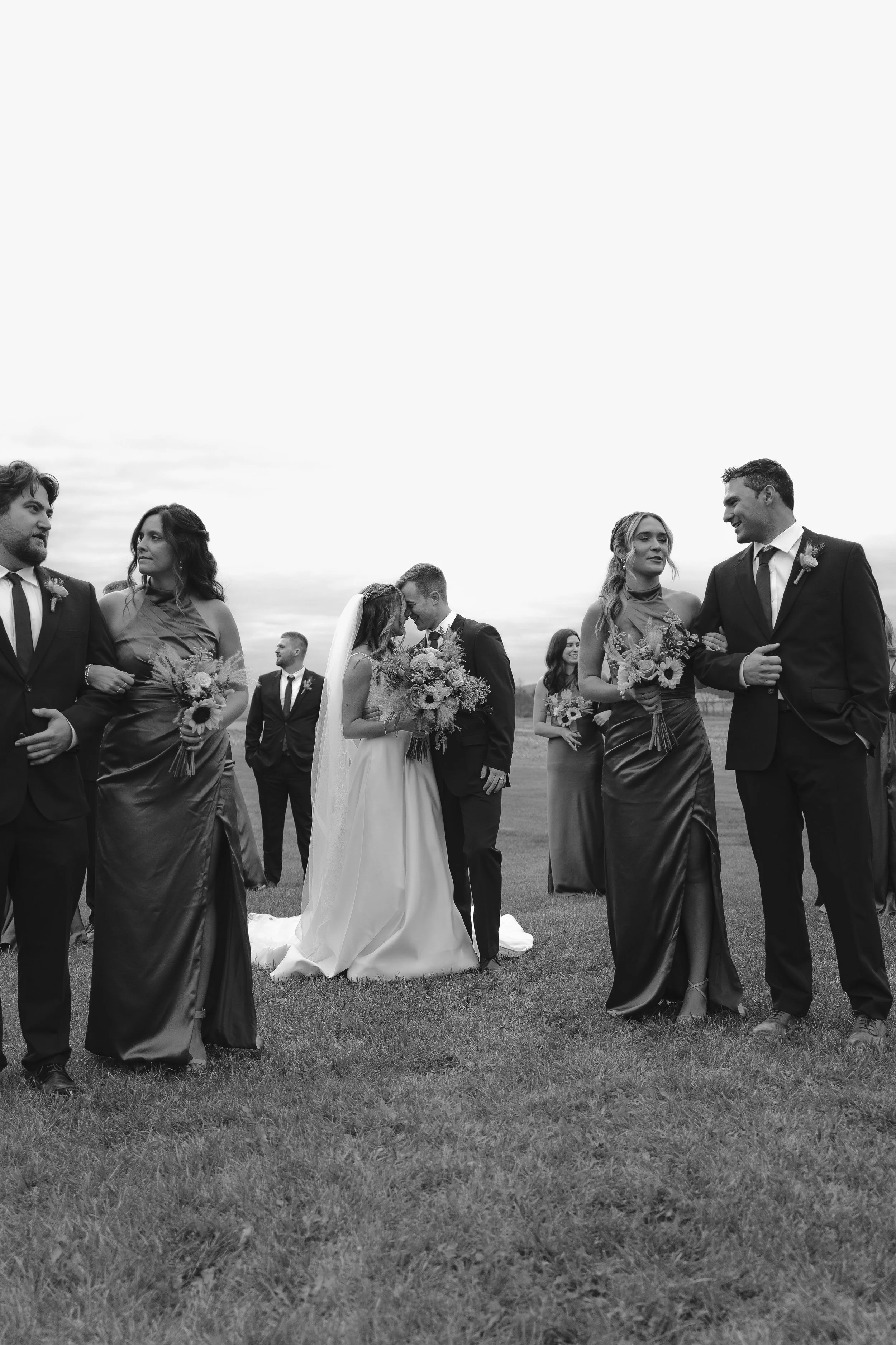 Black and white photo of a wedding ceremony outdoors, with brides and grooms exchanging vows while holding bouquets, surrounded by bridesmaids and groomsmen, on a grassy field with a cloudy sky in the background.