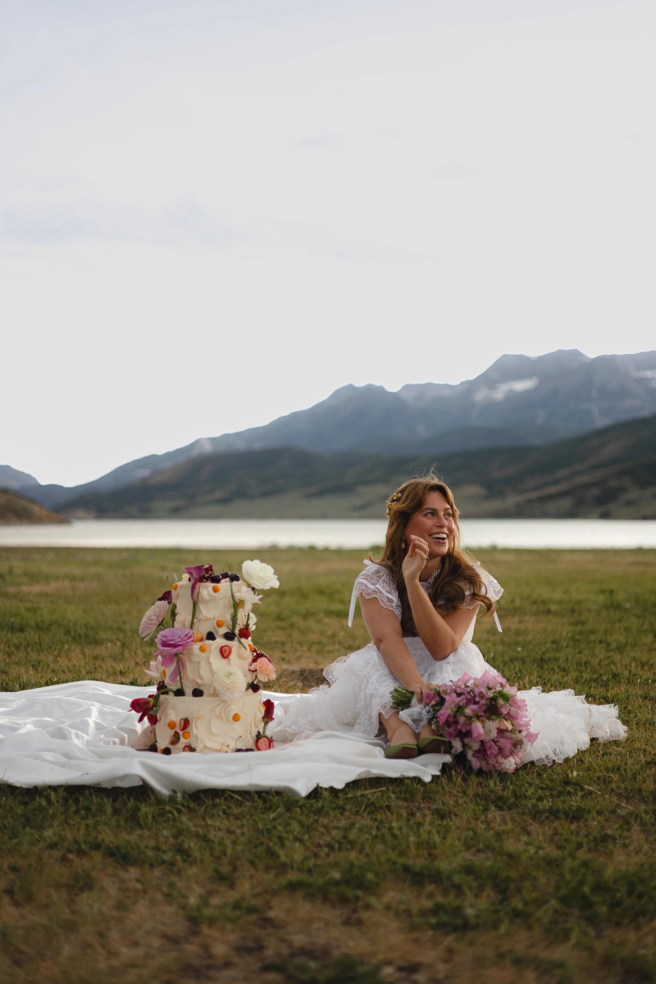 A woman in a white wedding dress sitting on a white cloth on the grass outdoors with a smiling expression. In front of her is a large, elaborate wedding cake decorated with flowers and berries. The woman has long, wavy hair and is surrounded by bouquets of pink and purple flowers. The background features a lake with mountains and cloudy sky.
