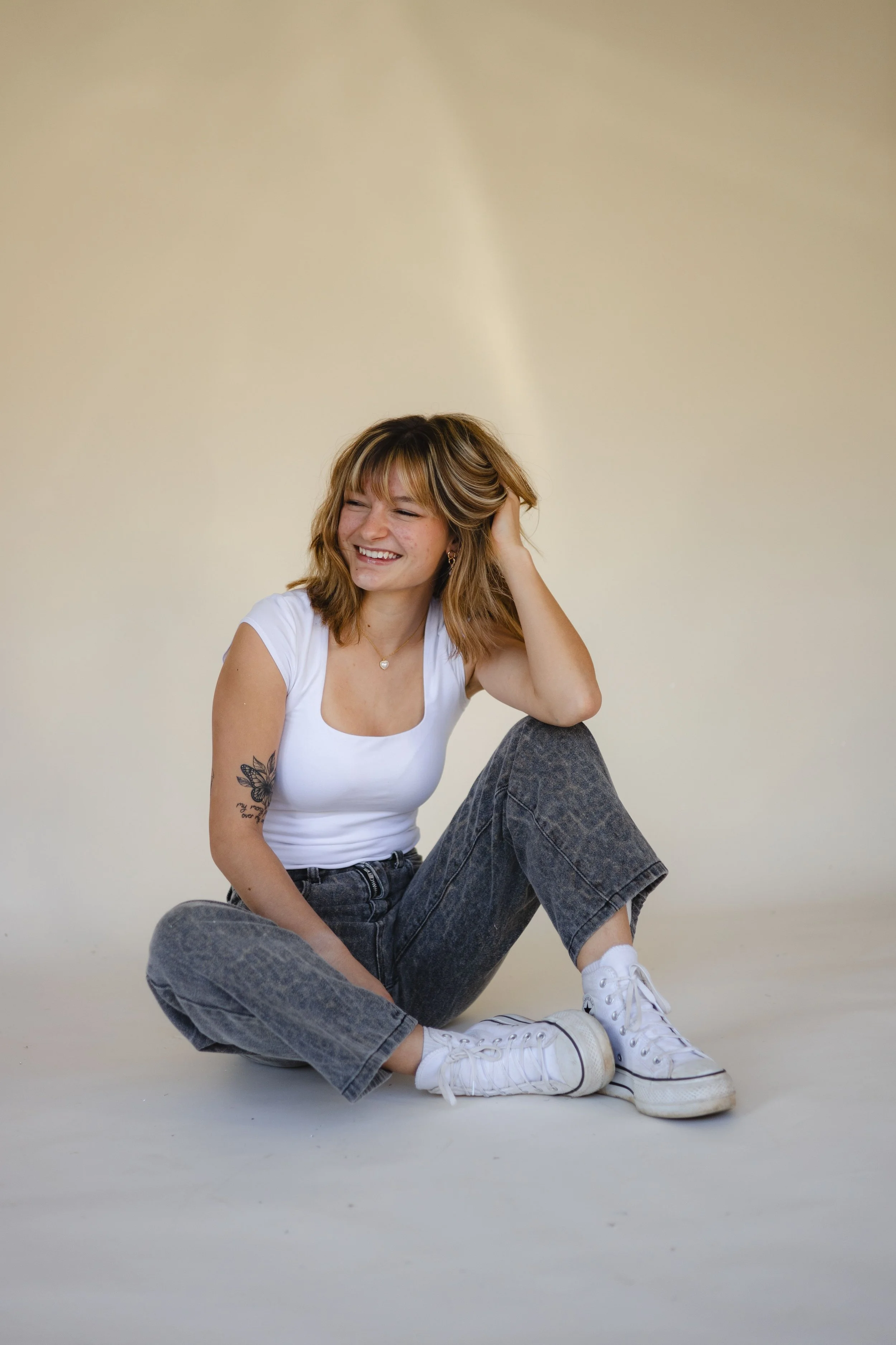 A smiling young woman sitting on the floor with her legs crossed, wearing a white t-shirt, gray jeans, and white sneakers, against a plain light-colored background.