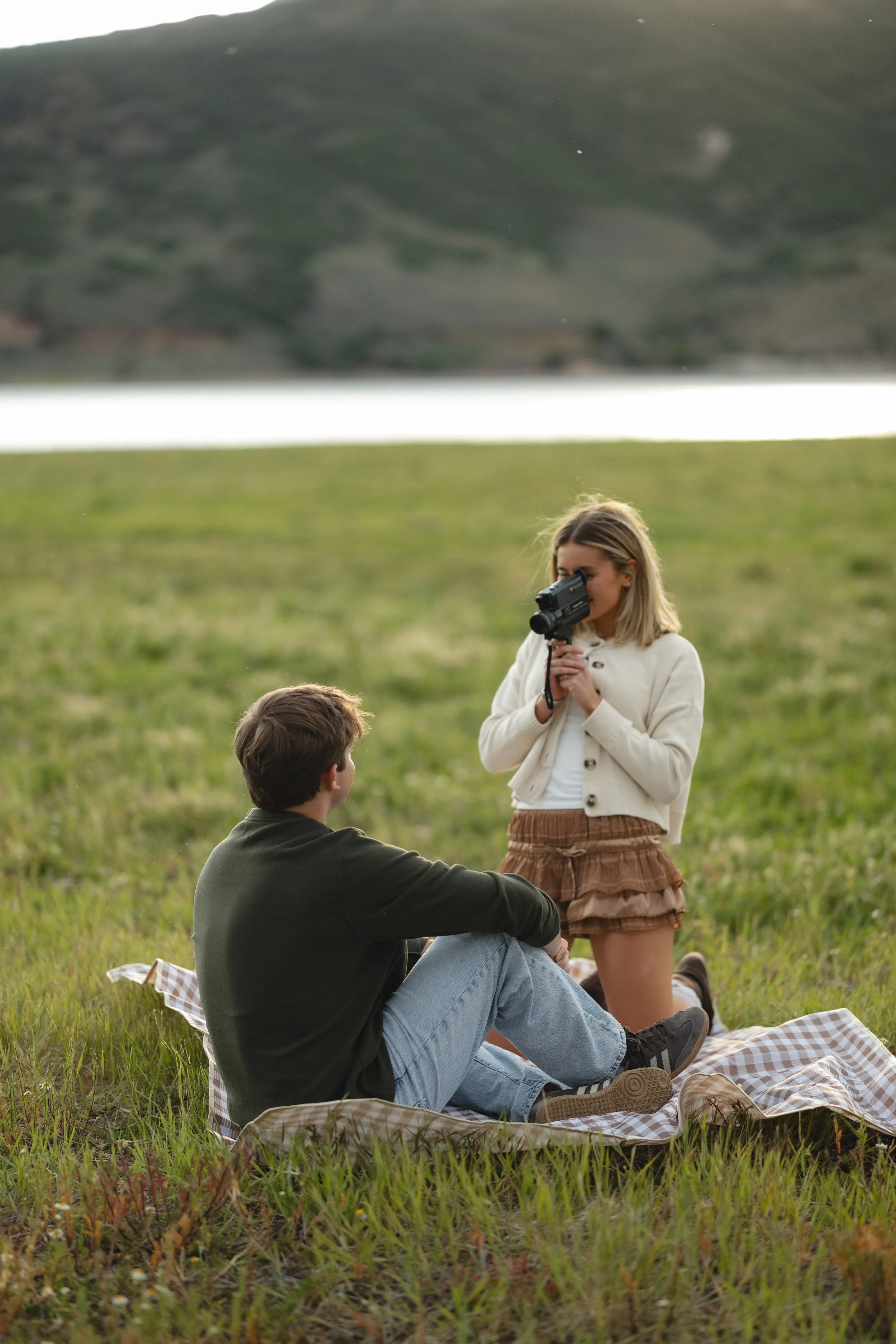 A woman holding a gun to her eye while standing in a grassy field, with a man sitting on a blanket and looking up at her, during sunset or late afternoon in an outdoor setting.
