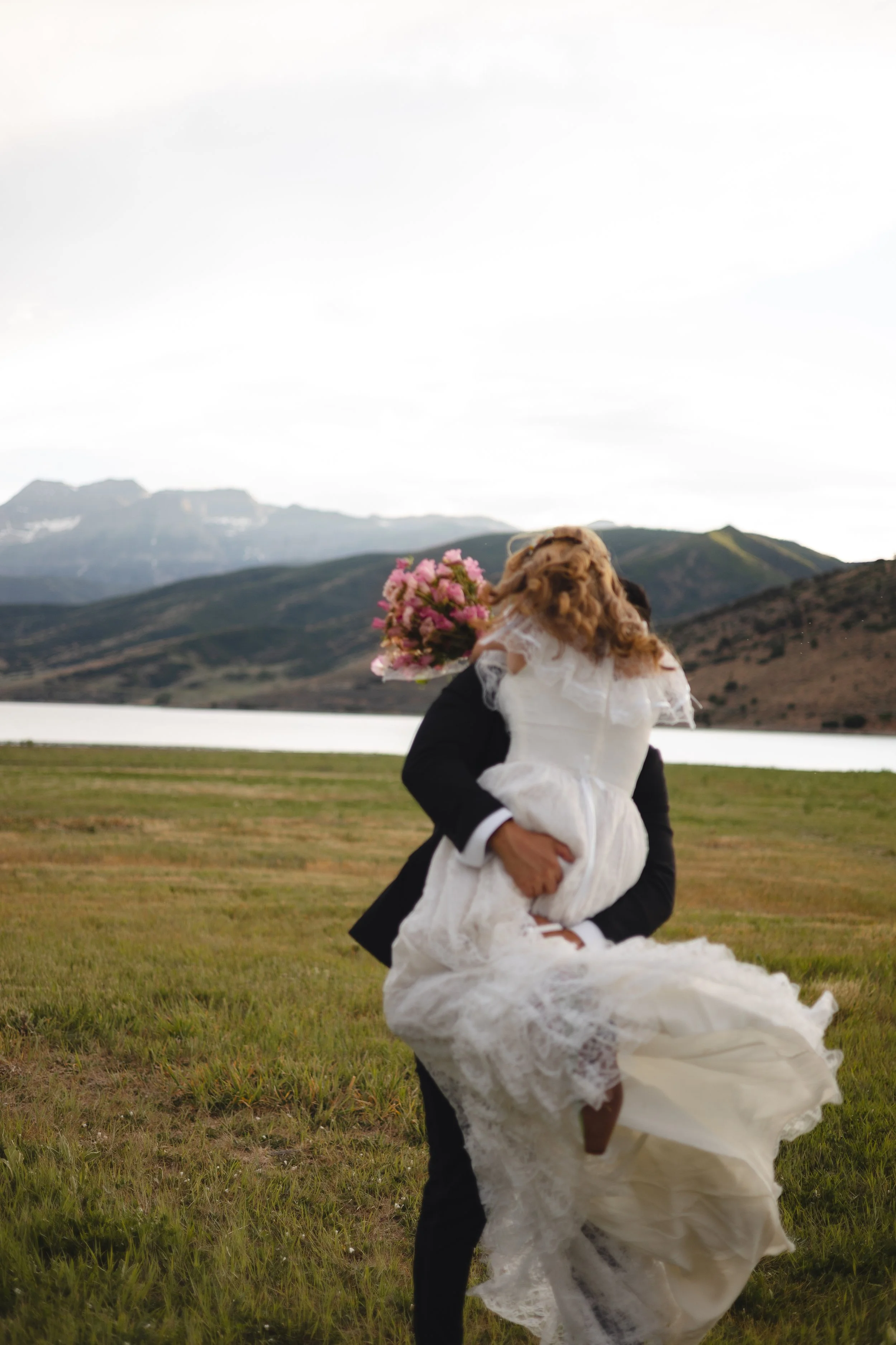A person in a black suit is holding a dog dressed in a white wedding dress, with a mountain and lake landscape in the background.