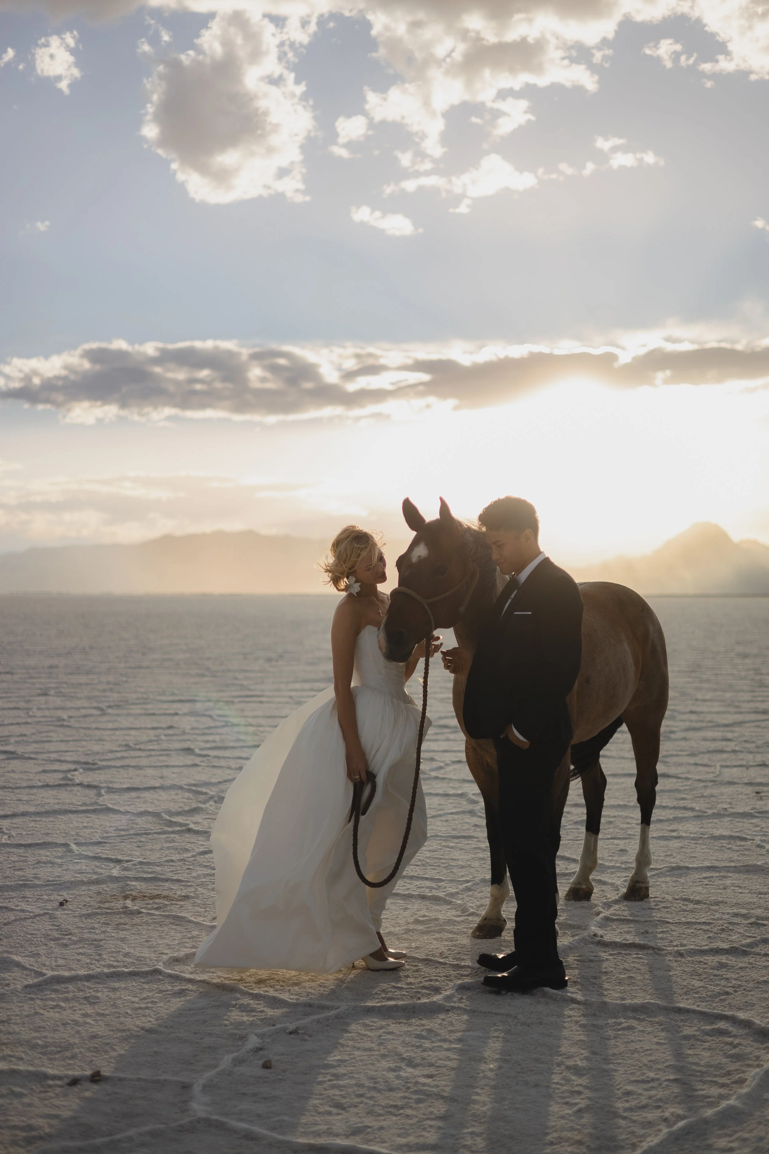 A couple dressed in wedding attire standing on a salt flat, holding a horse's reins, with a scenic sunset and mountains in the background.