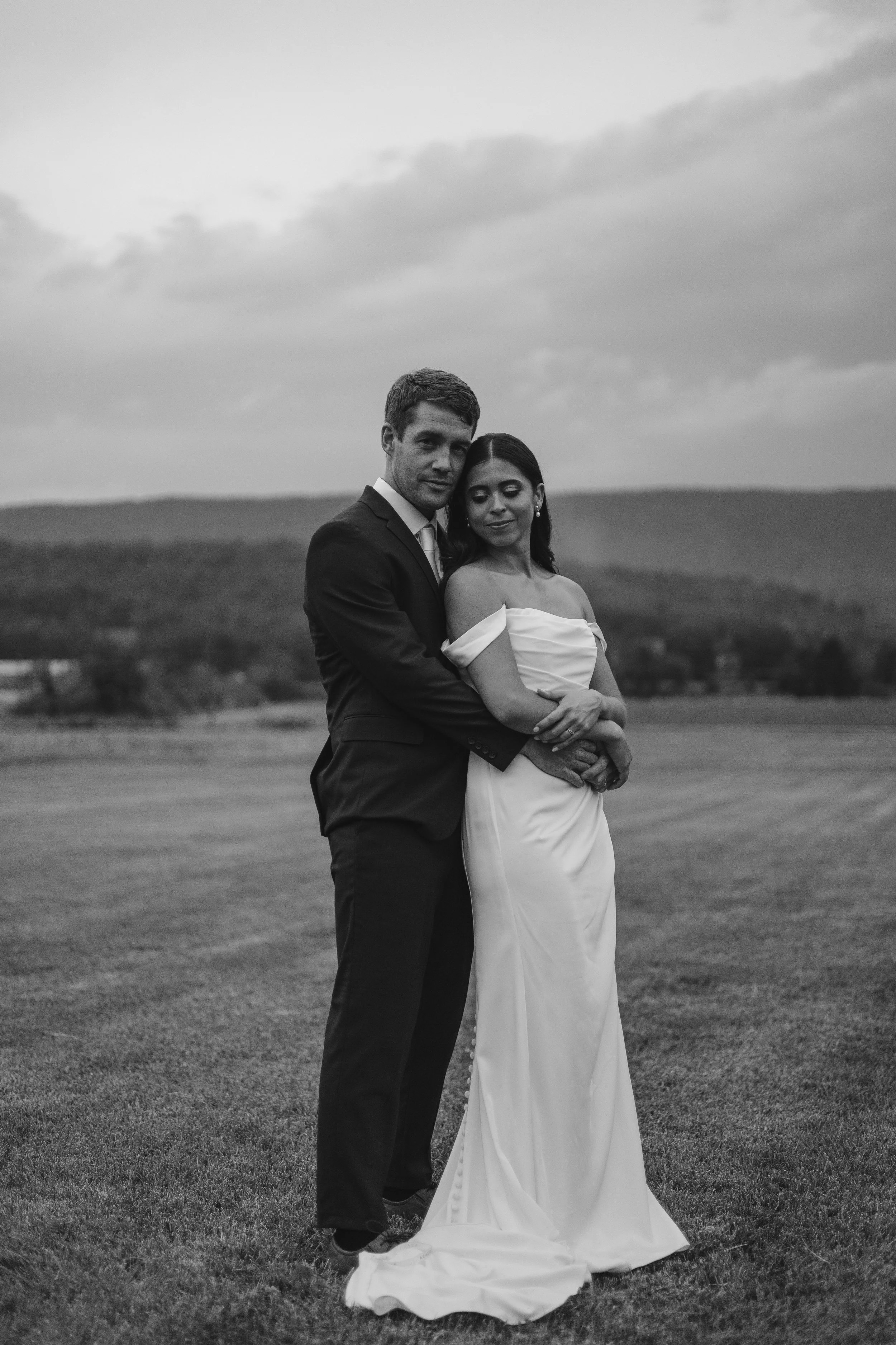 A black-and-white photo of a couple in wedding attire standing outdoors on a grassy field with hills in the background and cloudy sky above.