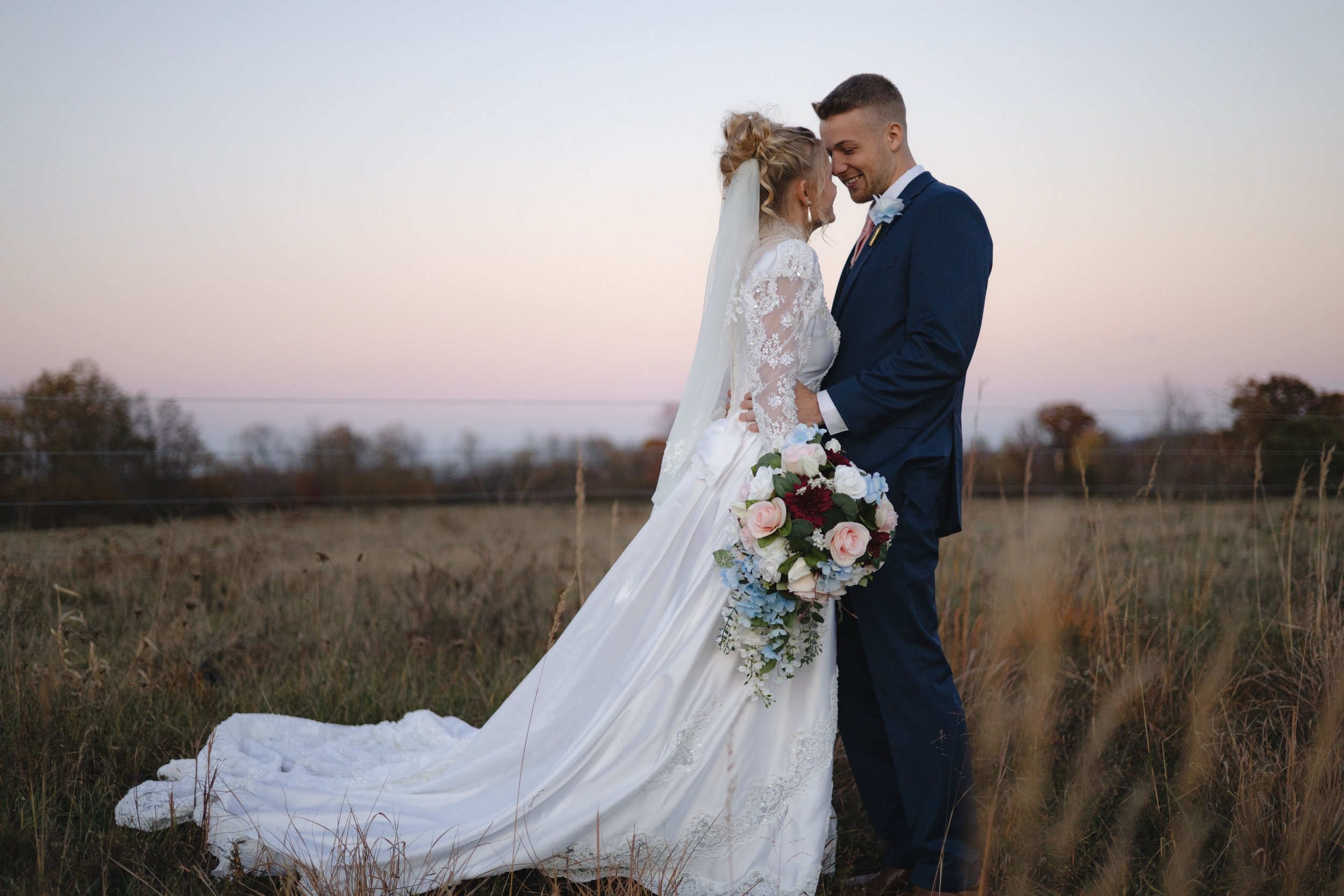 A bride and groom are standing close together outdoors during their wedding, smiling and touching foreheads at sunset, with a field and trees in the background.