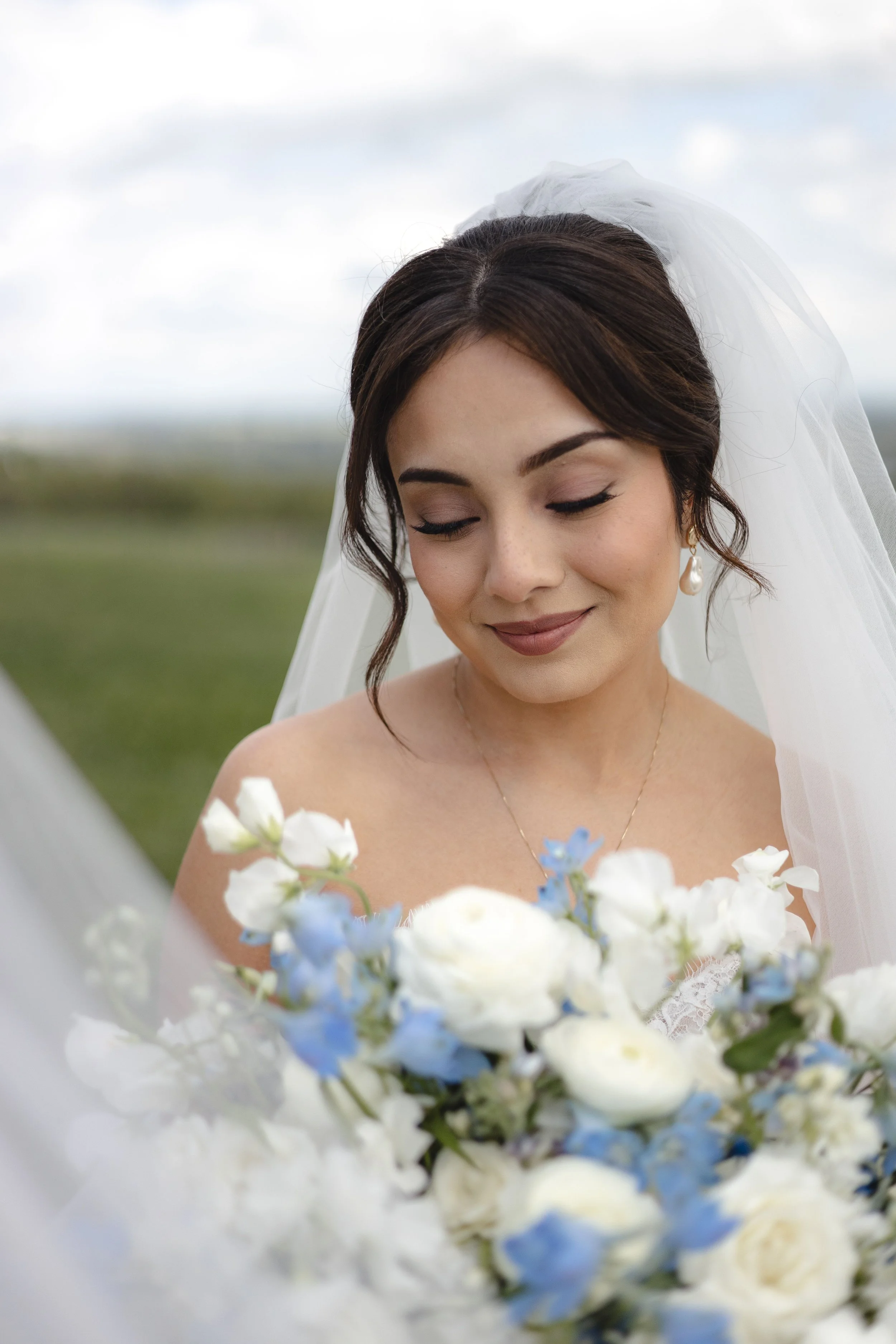 A bride with dark brown hair styled in loose waves, wearing a white veil, closed eyes, and a gentle smile, holding a bouquet of white and blue flowers, with a cloudy outdoor sky in the background.