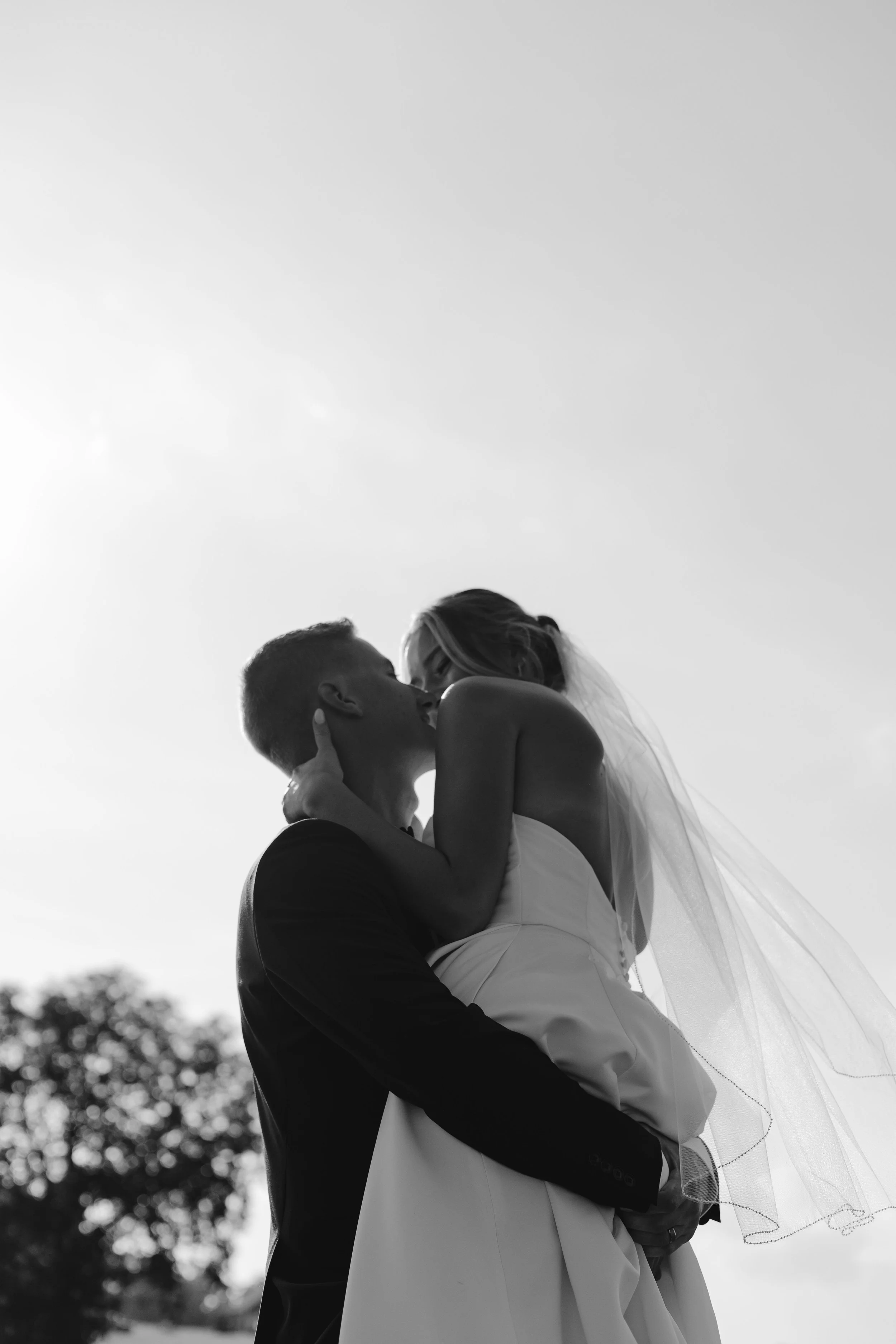 A black and white photo of a couple kissing outdoors during the day, with the groom holding the bride in his arms. The woman wears a wedding dress and veil, and the man is in a suit. The sky is clear with some trees in the background.