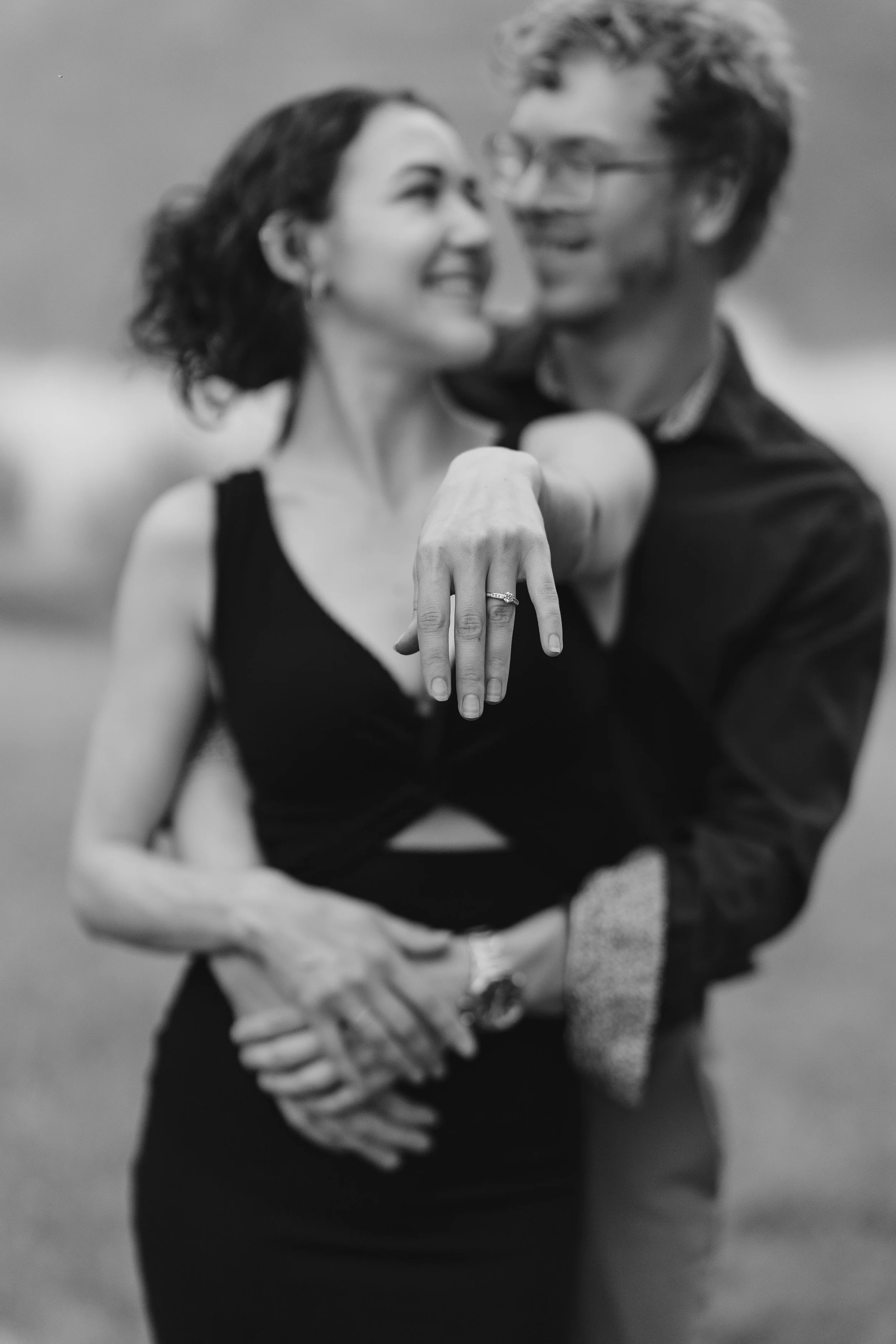 A couple celebrating their engagement, with the woman in the black dress reaching out her hand, showing off the ring that the man just placed there
