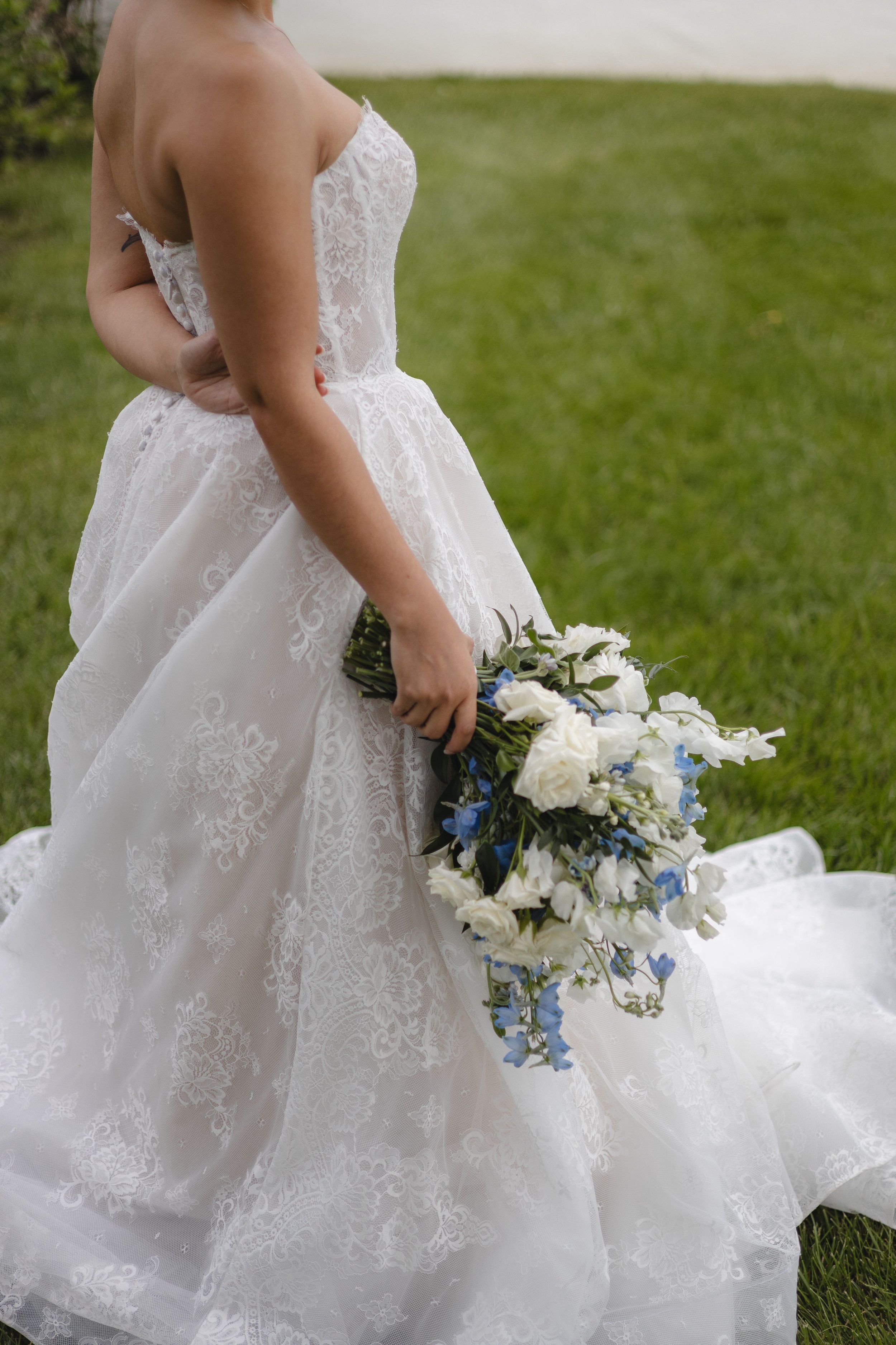 Close-up of a bride in a white floral lace wedding gown holding a cascading bouquet of white and blue flowers on a grassy outdoor setting.