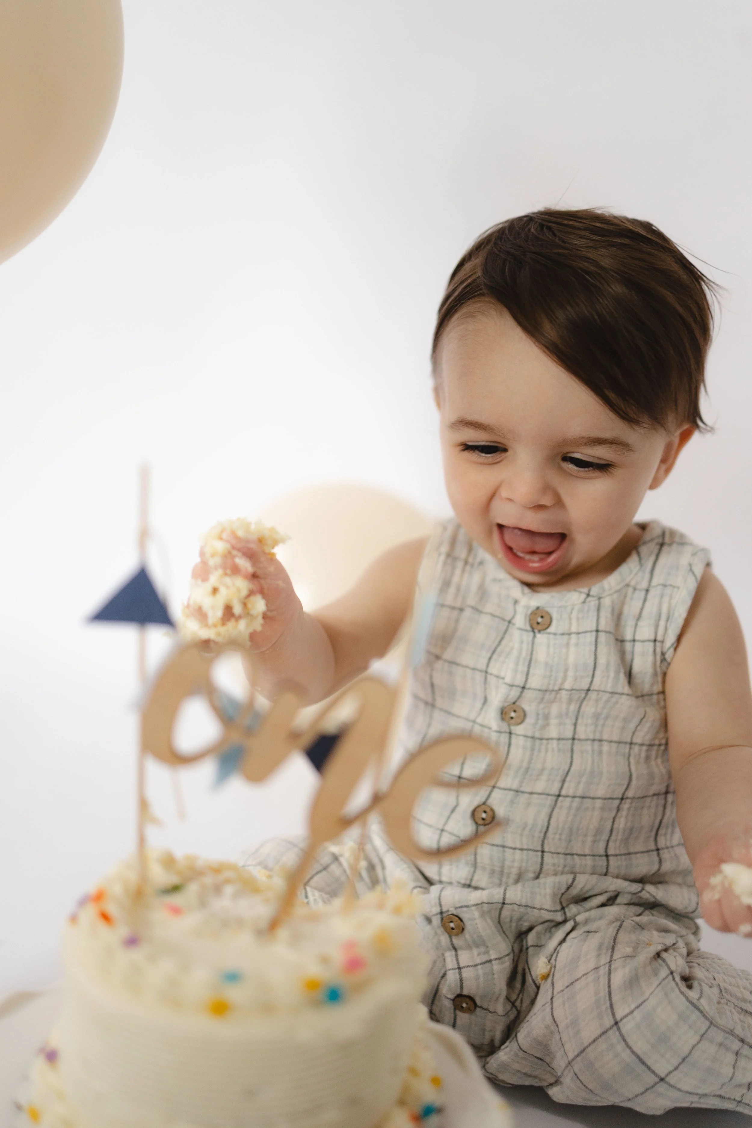 A young boy with brown hair and wearing a plaid romper is at a birthday celebration, looking at cupcakes with colorful sprinkles and decorations.