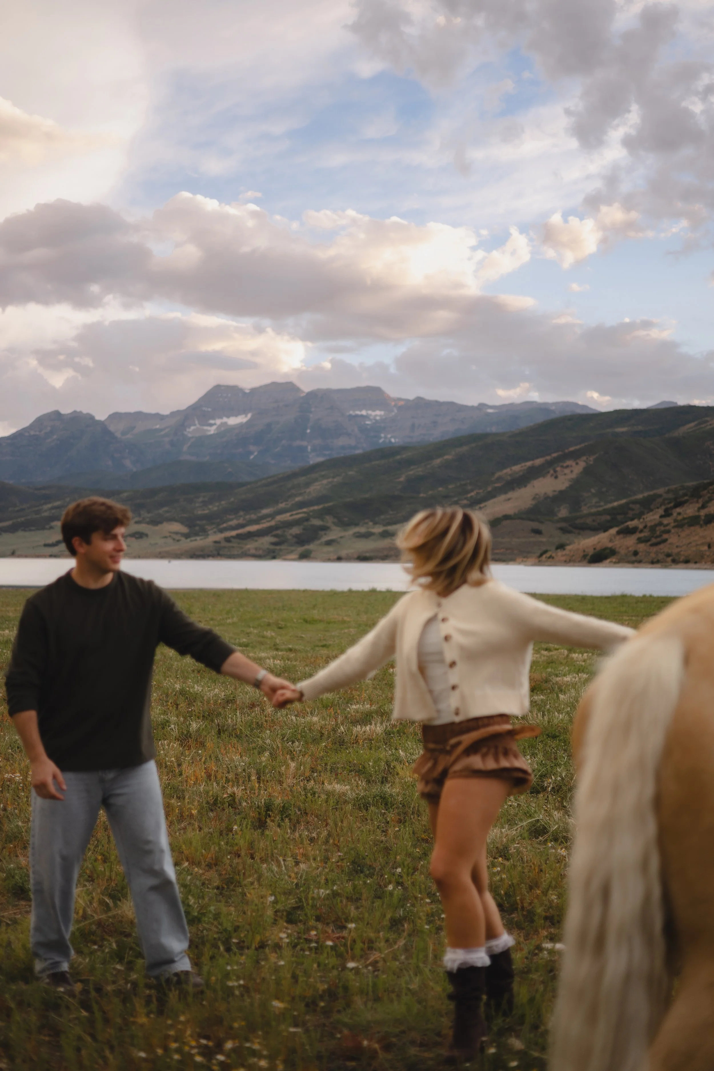 Two people holding hands in a grassy field with a lake, mountain range, and cloudy sky in the background.