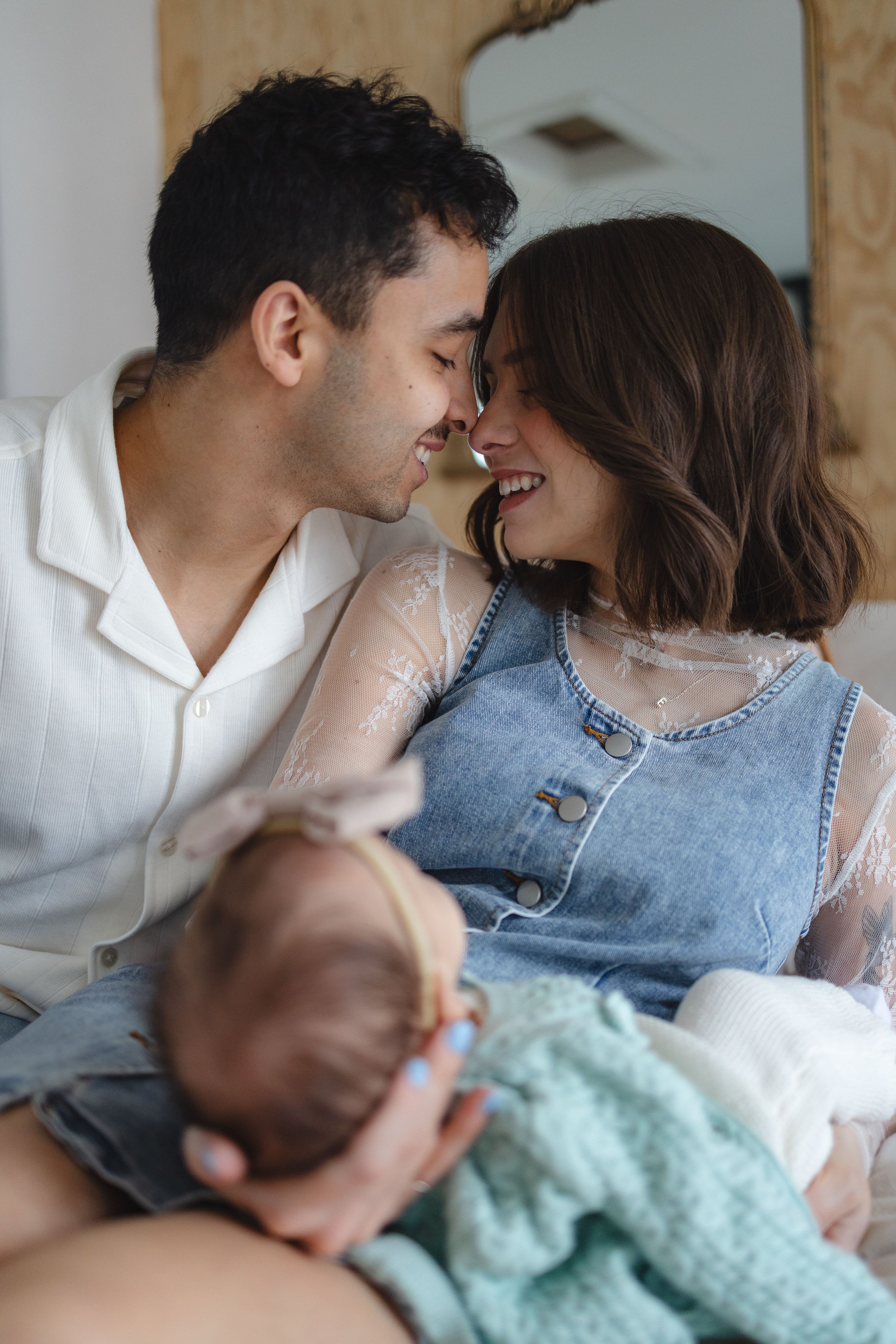 A couple smiling with foreheads touching, holding a newborn baby, with a mirror and a wooden frame in the background.