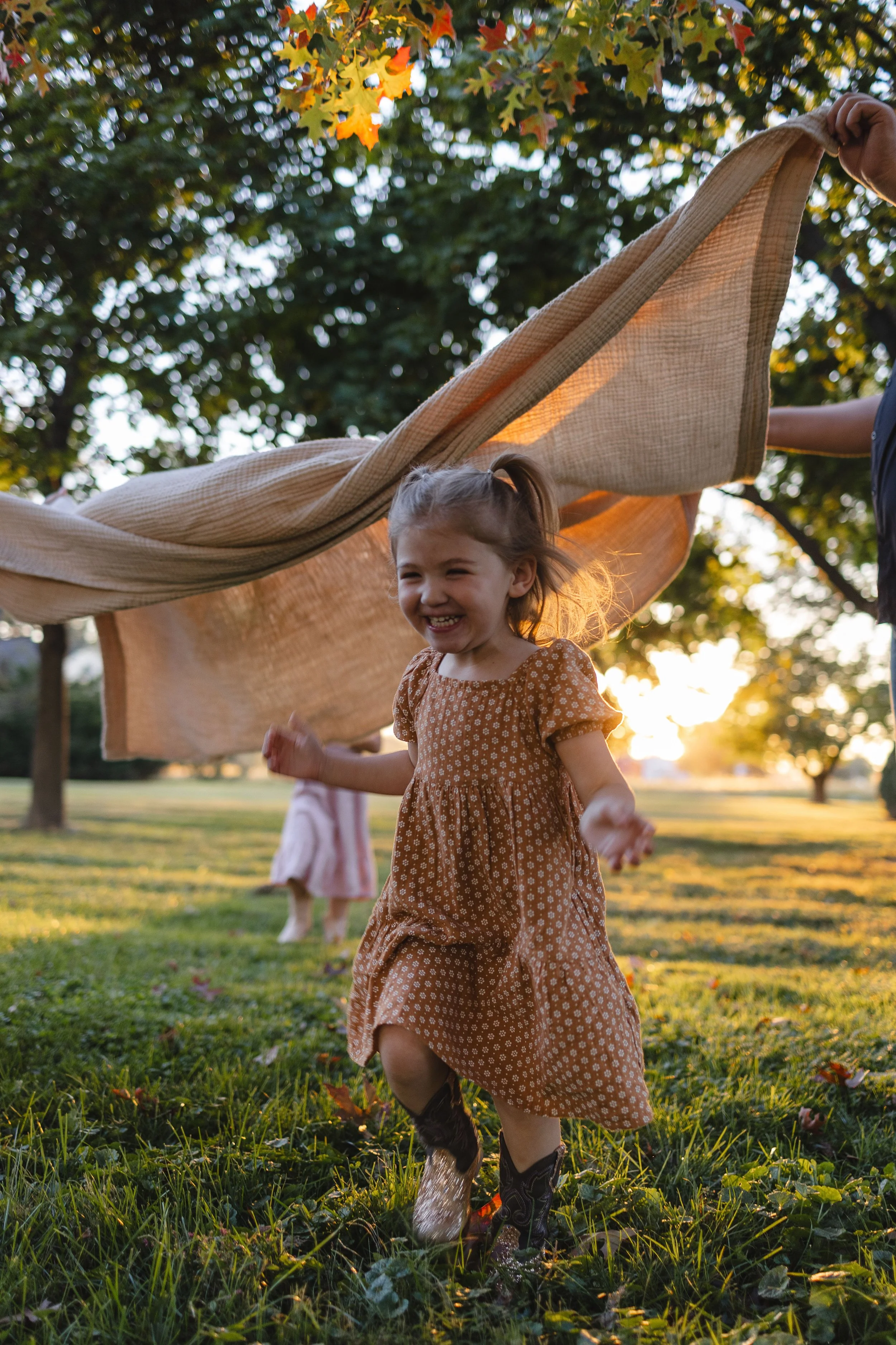 A young girl with a ponytail, wearing a brown dress with white polka dots and cowboy boots, is smiling and running on grass during golden hour in a park with trees. Another girl in a pink dress is in the background, and a person is holding a large beige cloth or blanket above the girl.