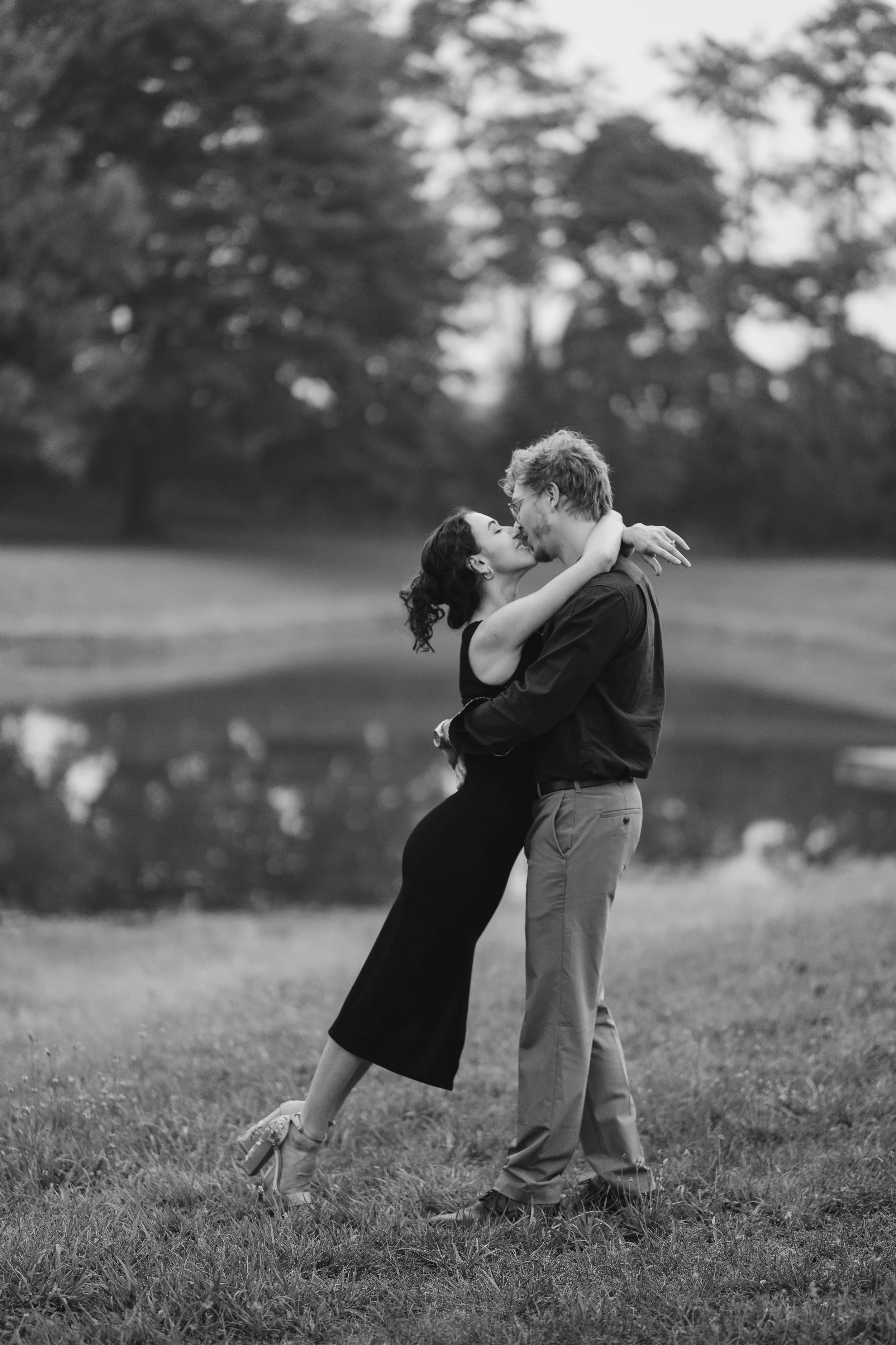 Girl in a black dress leans into a boy wearing a button down shirt, as she drapes her hands over his neck and touches her lips to his, with a big smile while doing it all in front of a small lake