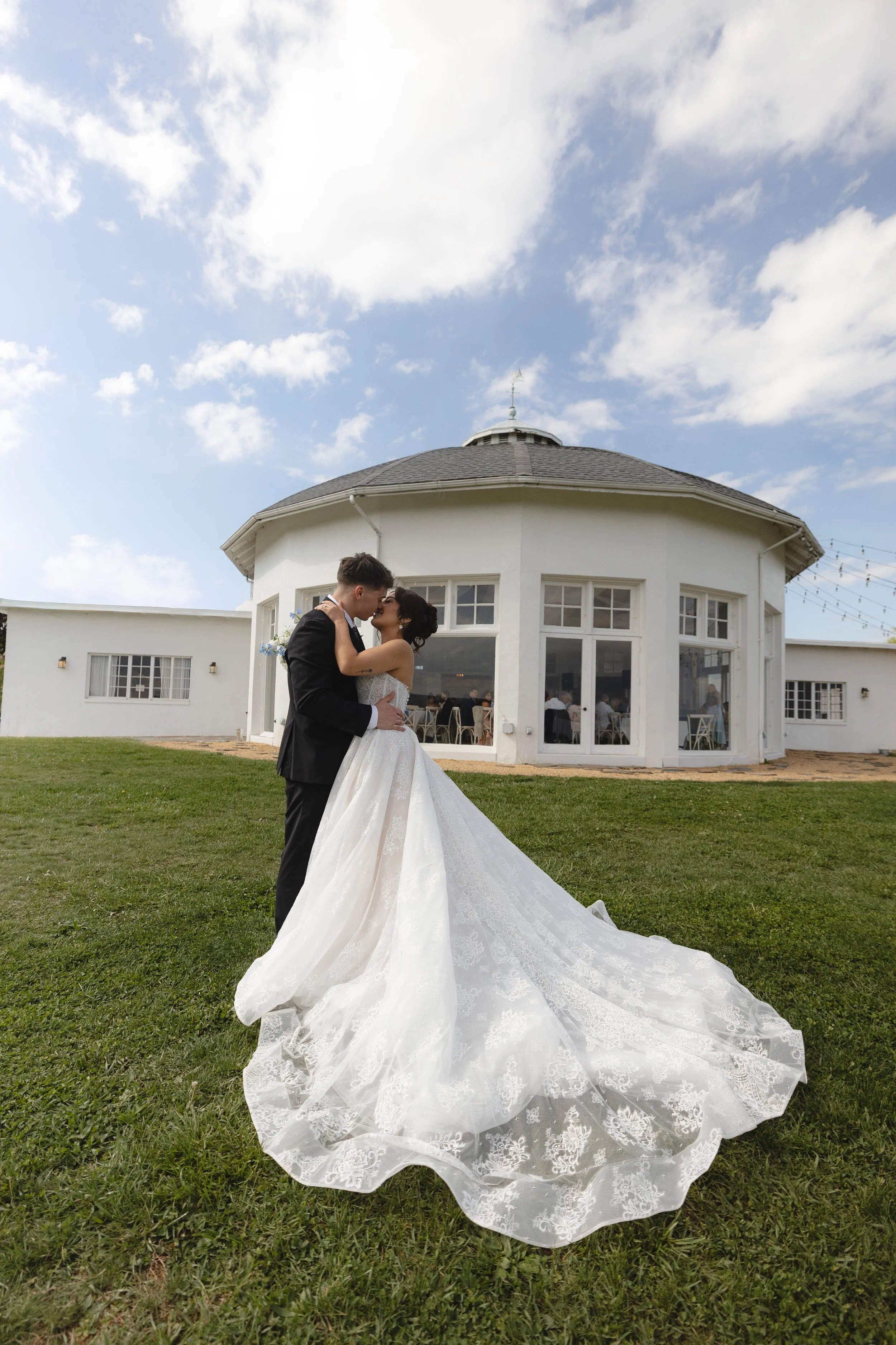 A bride and groom sharing a kiss outside a white, round building with large windows on a grassy lawn under a partly cloudy sky.