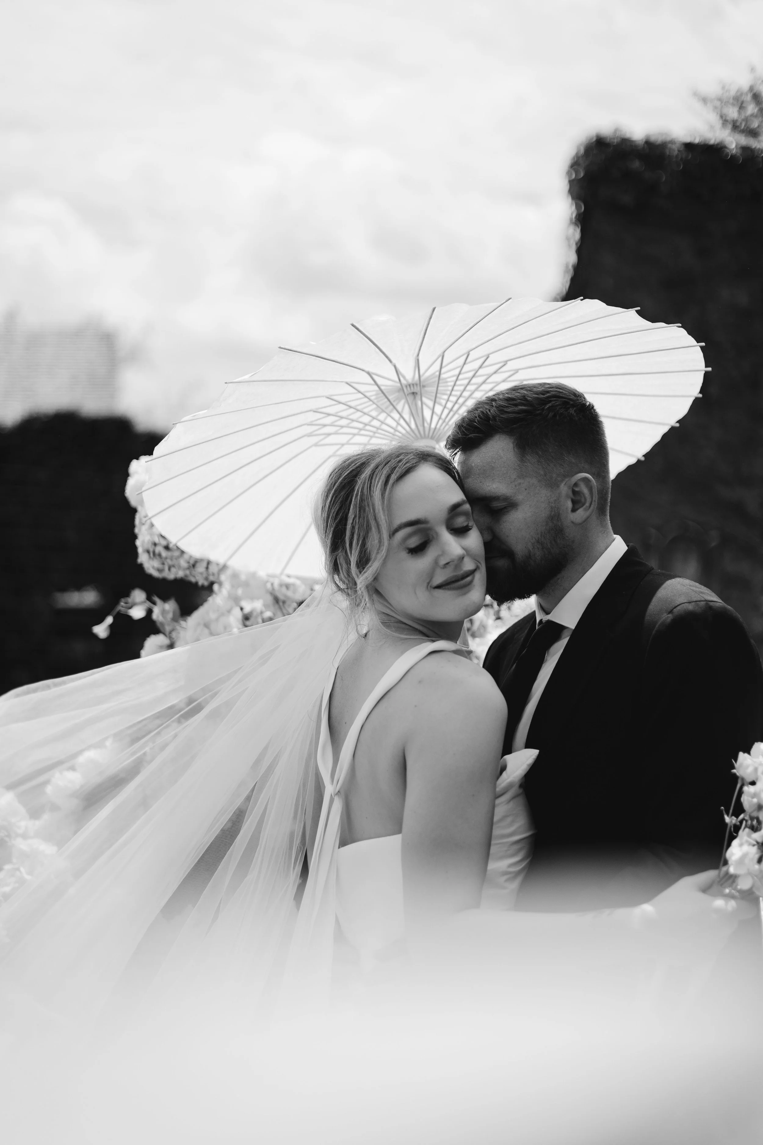 Black and white photograph of a bride and groom embracing under a parasol, with the bride's veil flowing in the wind, set outdoors with trees and cloudy sky in the background.