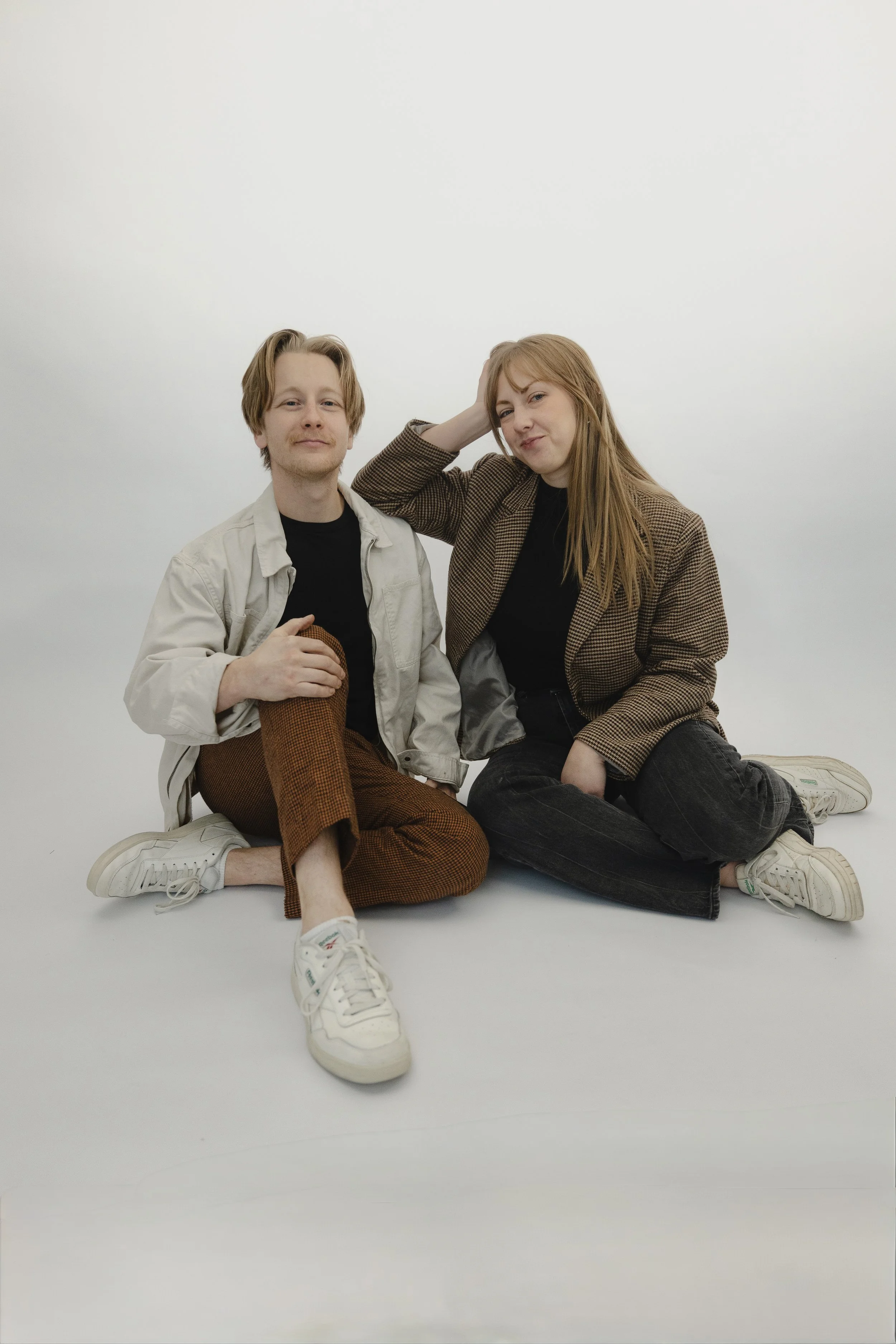 A young man and woman sitting on the floor against a plain white background. The man has light brown hair, is wearing a light-colored jacket, black shirt, brown plaid pants, and white sneakers. The woman has long red hair, is wearing a brown checkered blazer, black shirt, dark jeans, and white sneakers. They are posing casually, with the woman touching her head with one hand and the man holding a baseball glove.