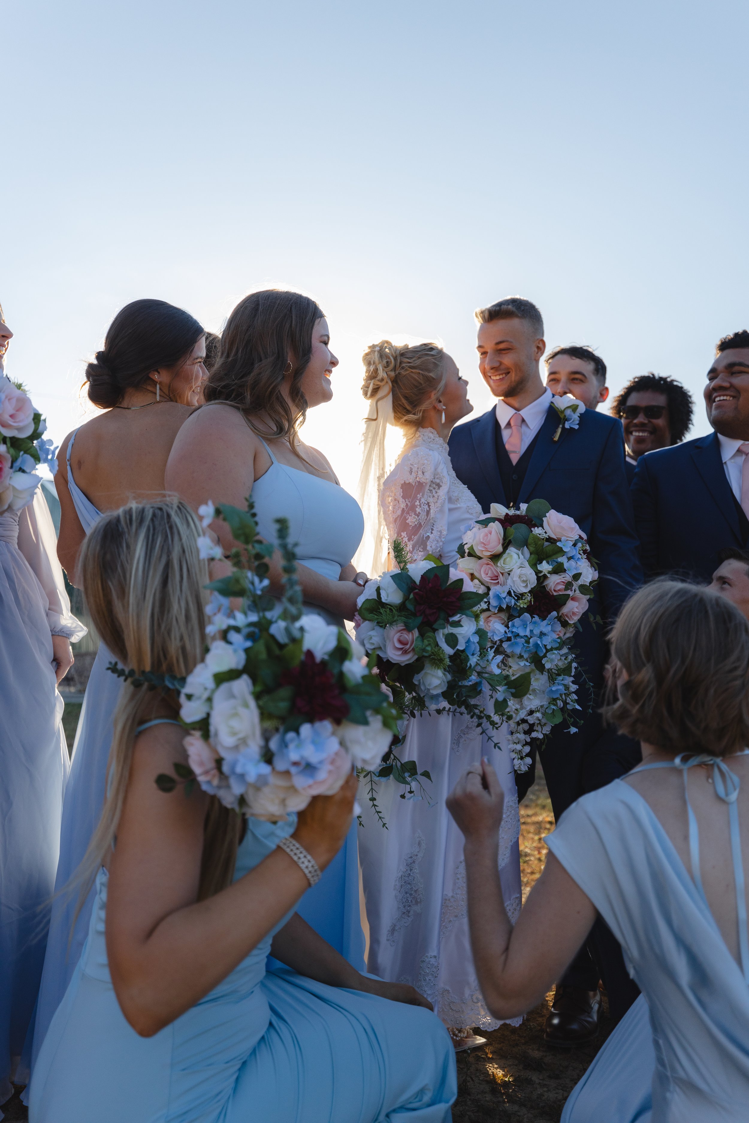 A group of people gathered outdoors for a wedding, with a bride and groom at the center, smiling and exchanging glances, surrounded by bridesmaids and guests holding flowers, all dressed in formal attire on a sunny day.
