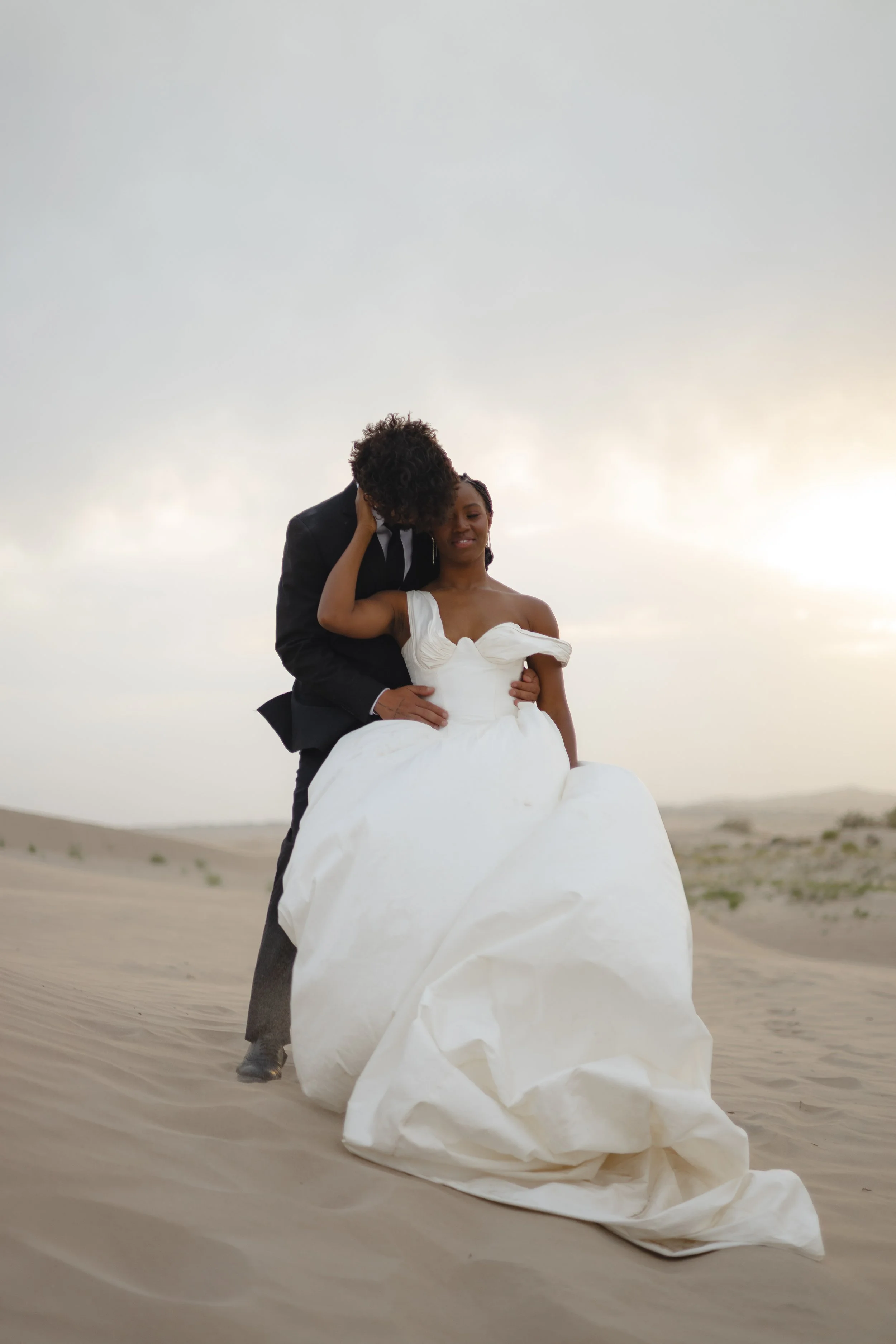 A couple in wedding attire standing on a sandy beach at sunset, with the bride wearing an off-shoulder white wedding gown and the groom in a black suit, sharing an intimate moment.