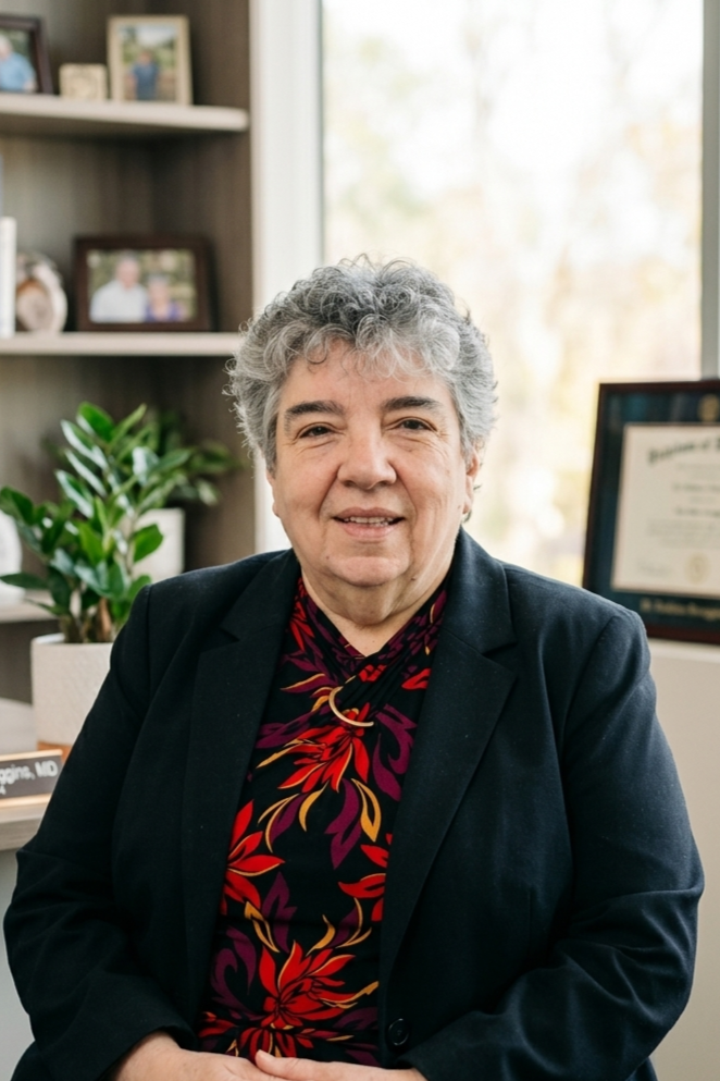 An older woman with short gray curly hair wearing a black blazer and a dark floral blouse, sitting in an office with framed certificates and photos in the background.