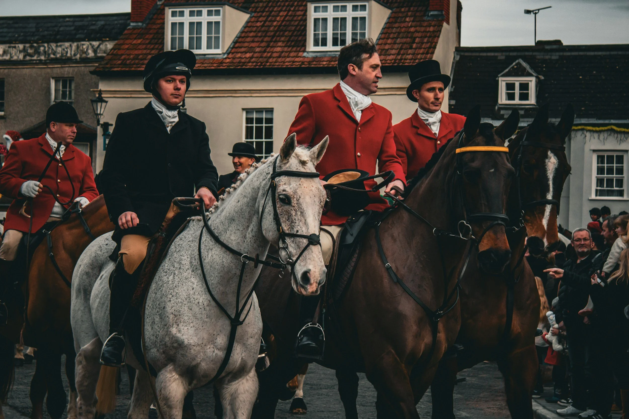 Group of people riding horses in a town square, dressed in historical clothing with some wearing red coats and black hats, with buildings and spectators in the background.