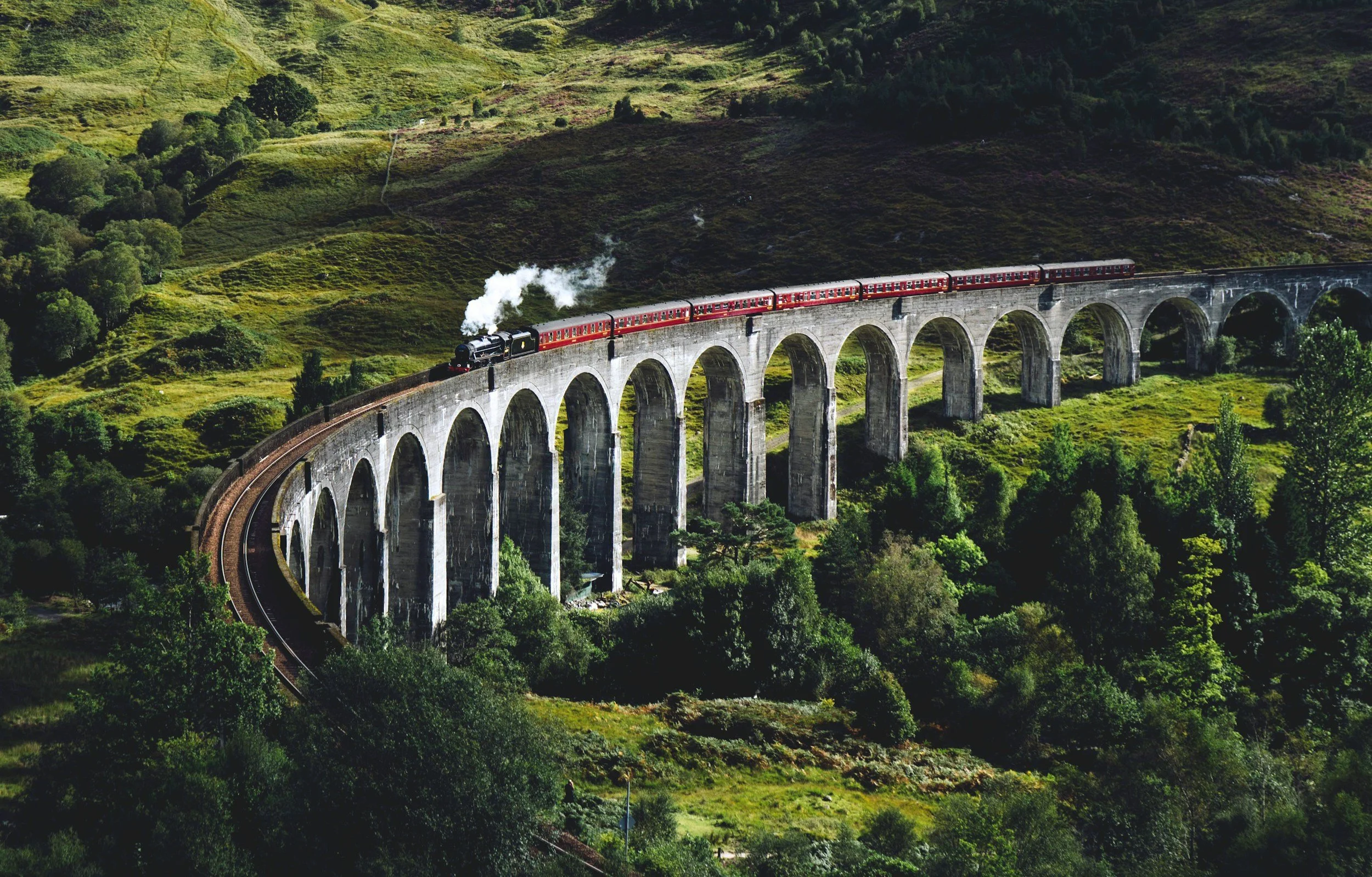 A steam train traveling across a high arched stone bridge in a green mountainous landscape.