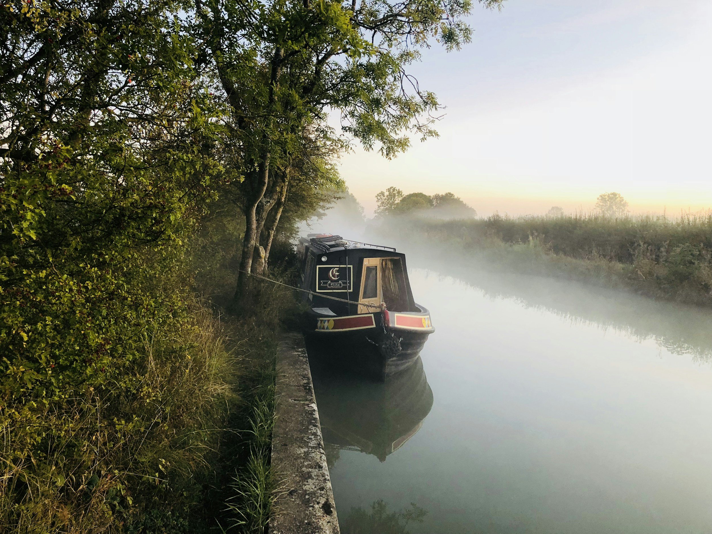 A boat docked along a calm, misty river or canal in a natural setting with trees and foliage.