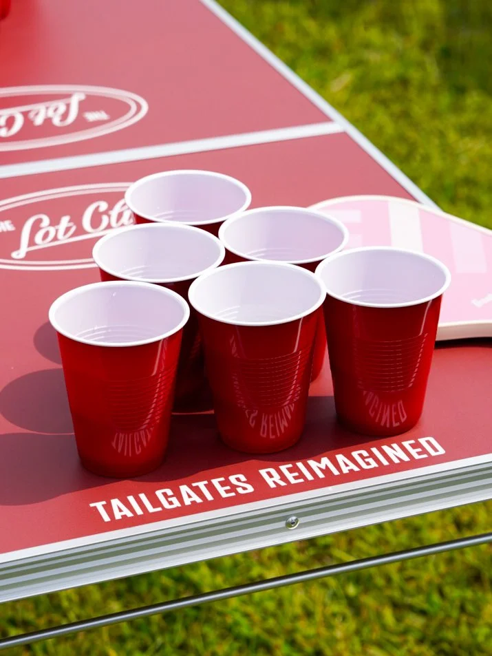 Six red plastic cups are arranged on a table with a red and white design, with the phrase 'TAILGATES REIMAGINED' visible, outdoors on grass.
