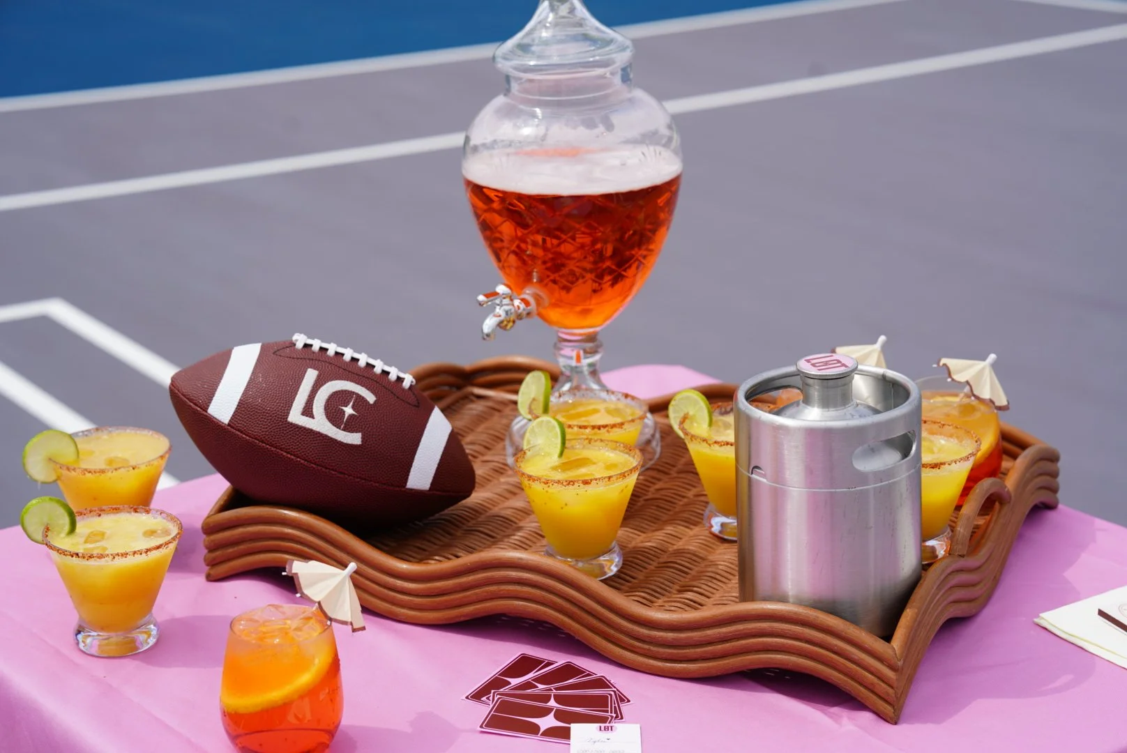 A wicker tray on a pink tablecloth with yellow cocktails garnished with lime slices, a large glass dispenser with amber-colored beverage, a football, and a metal container with a tap. There are small cocktail umbrellas, a stack of playing cards, and a fuzzy background of tennis courts.