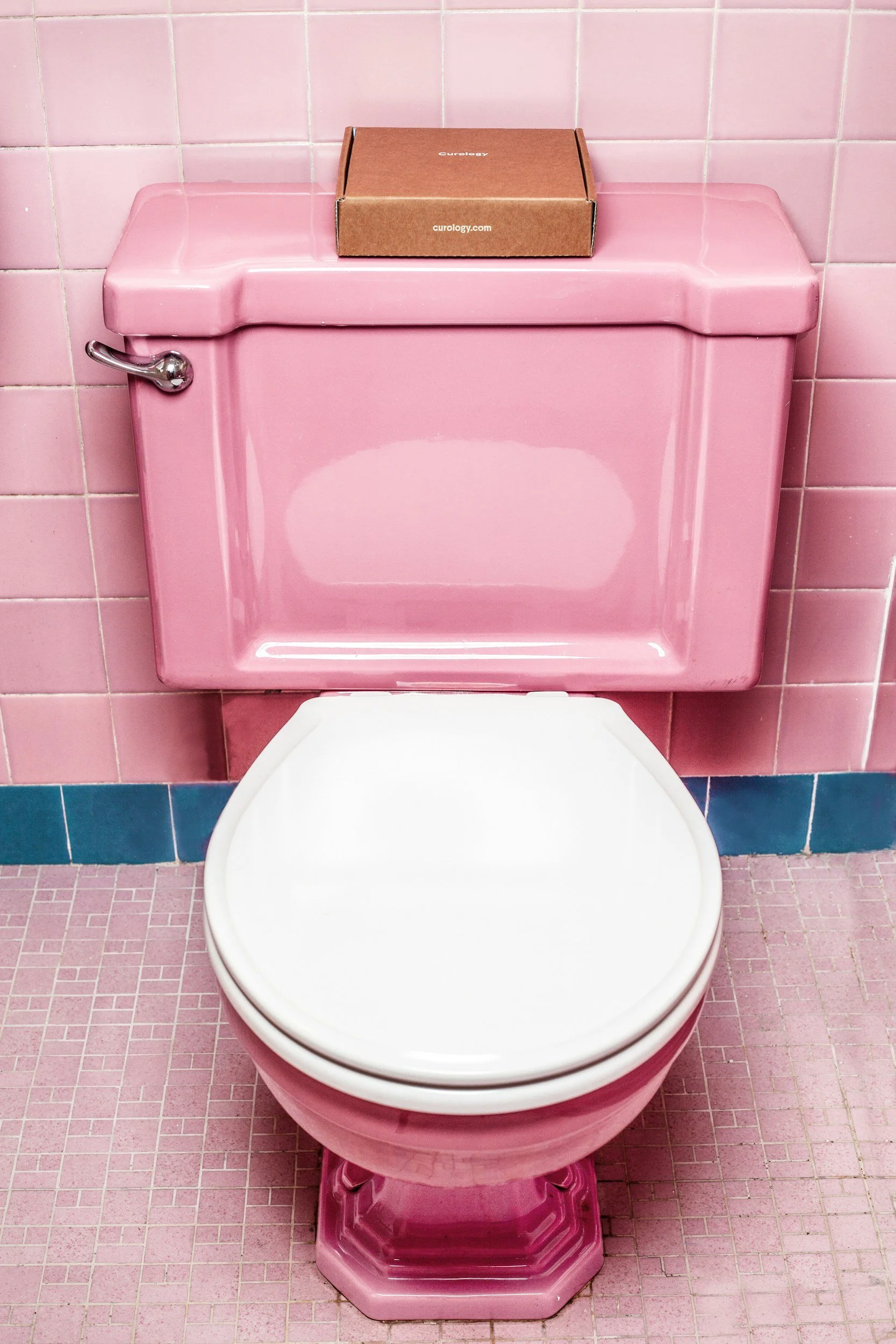 A pink toilet with a pink tiled bathroom wall and floor. There is a cardboard box labeled 'curology' on top of the tank.