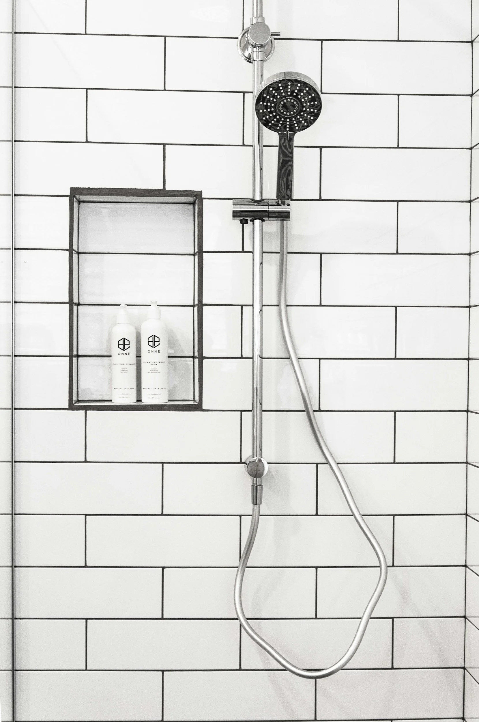 A showerhead with a hose attached, mounted on a wall with white rectangular tiles with black grout lines. There is a built-in shelf holding two bottles of toiletries labeled 'ONNE'.