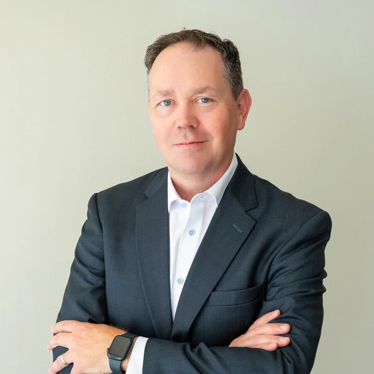 Portrait of Dr. Dan Young in a dark suit with a white shirt, standing against a light-colored background, looking confidently at the camera with arms crossed.