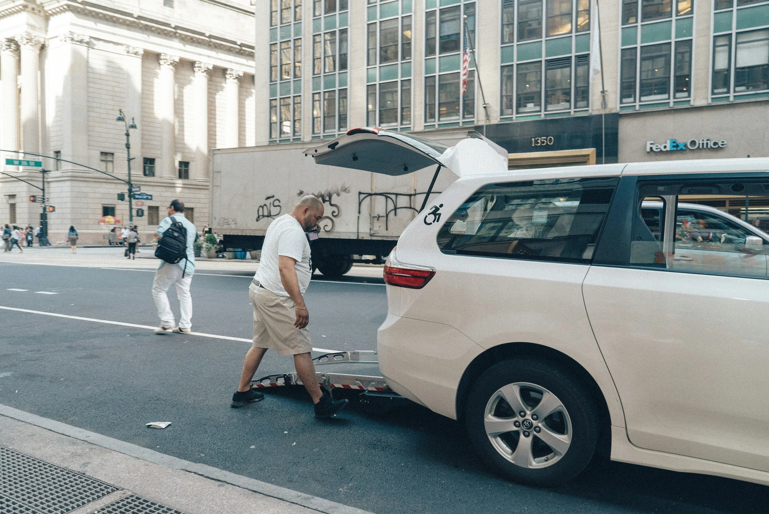 A man in a white shirt and beige shorts is opening the trunk of a white car parked on the city street. There are pedestrians walking across the street and tall buildings in the background, including a FedEx Office and a street sign reading 'E 34th St'.