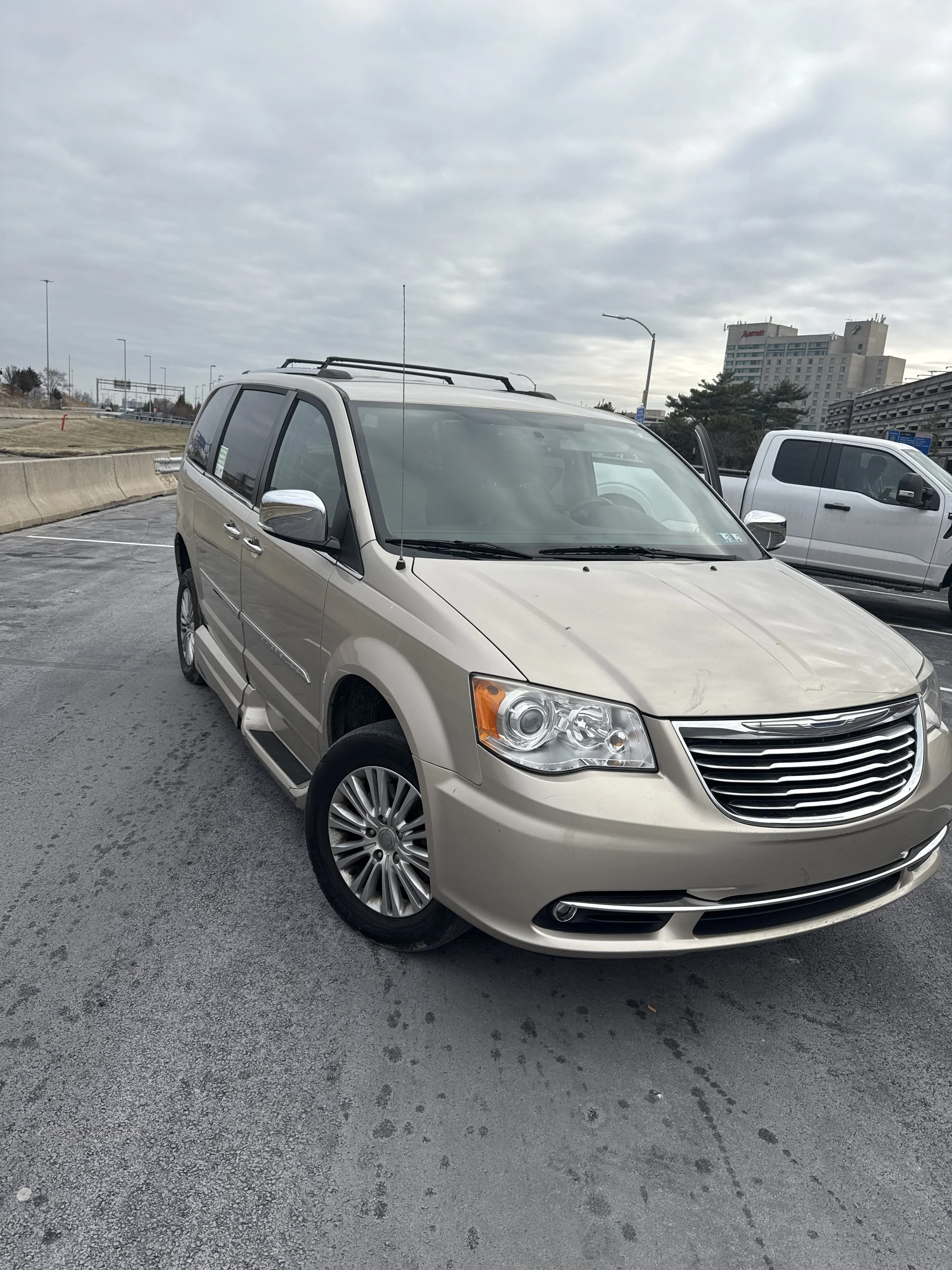 A beige minivan parked in a parking lot with another silver pickup truck nearby. Overcast sky with a cityscape in the background, including a Marriott hotel.