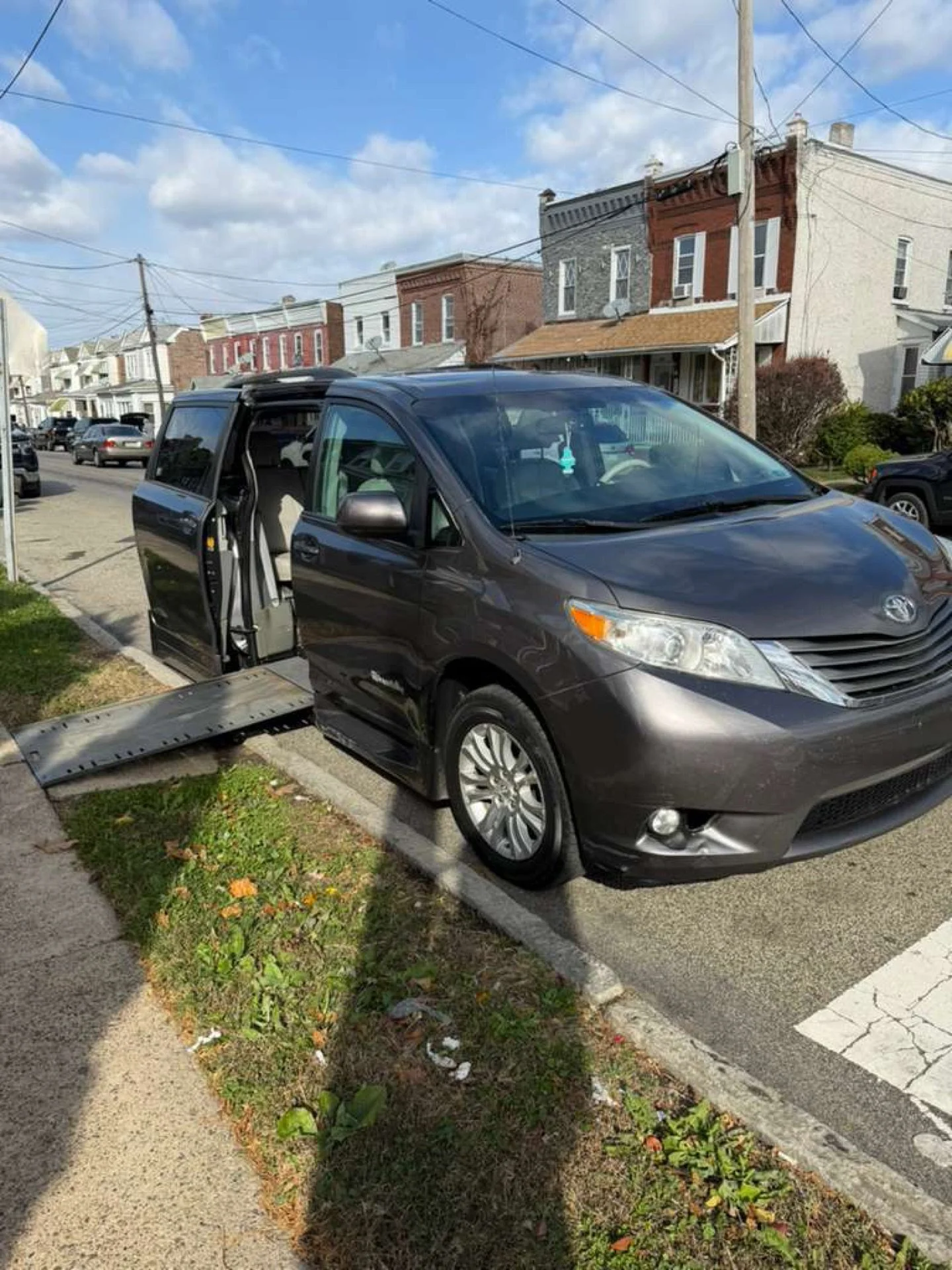 Black minivan parked partially on the sidewalk with its sliding door open, a wheelchair lift extended under the door, in a residential neighborhood with houses, trees, and power lines.