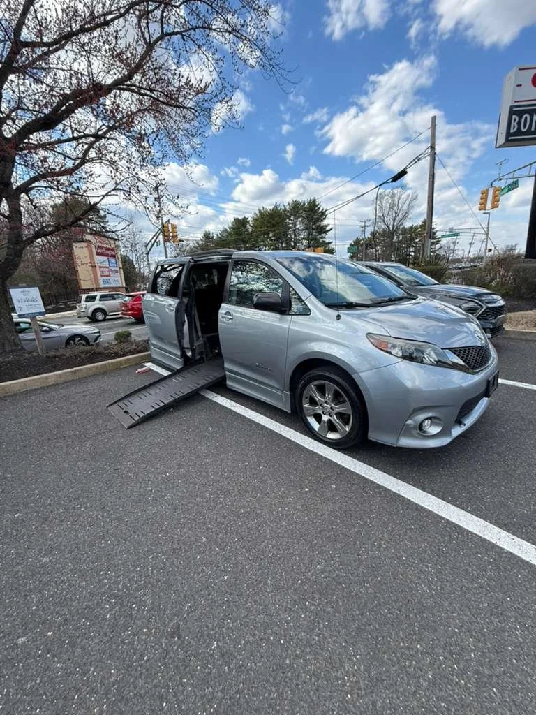Silver minivan with a wheelchair ramp deployed in a parking lot on a partly cloudy day.