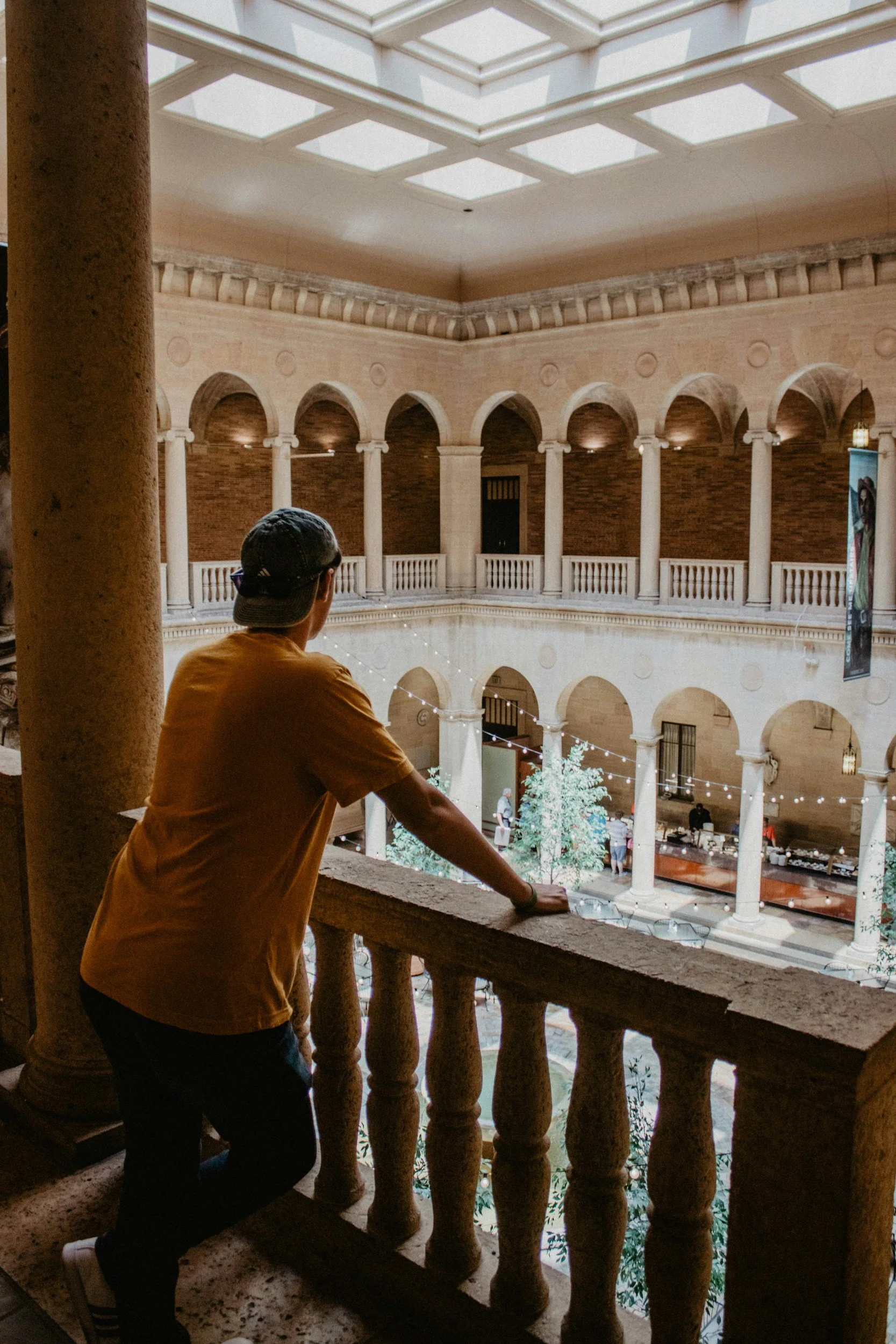 A person in a yellow shirt and cap leaning on a stone railing on a balcony, looking down at a courtyard decorated with  string lights inside a building with arched openings and columns.
