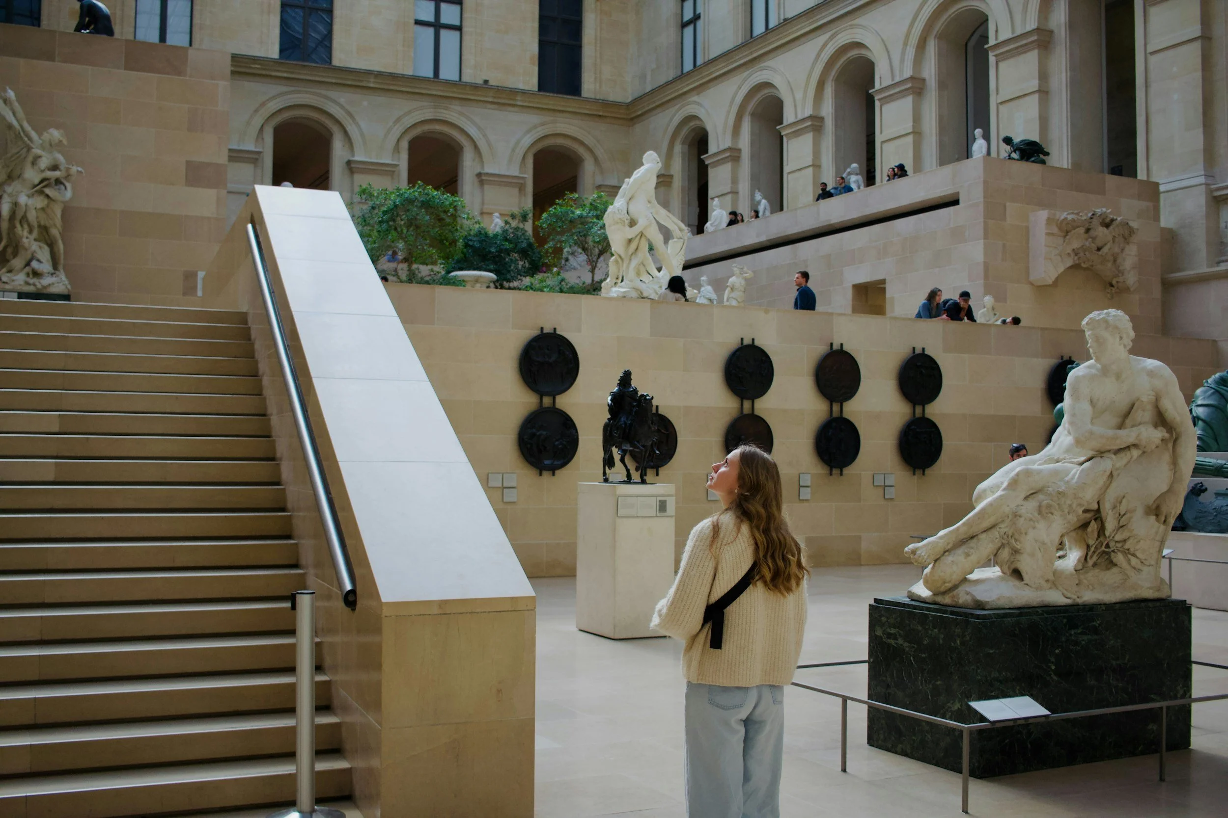 Women observing sculptures in an art museum interior with stairs, large arched windows, beige walls, and various statues on display.