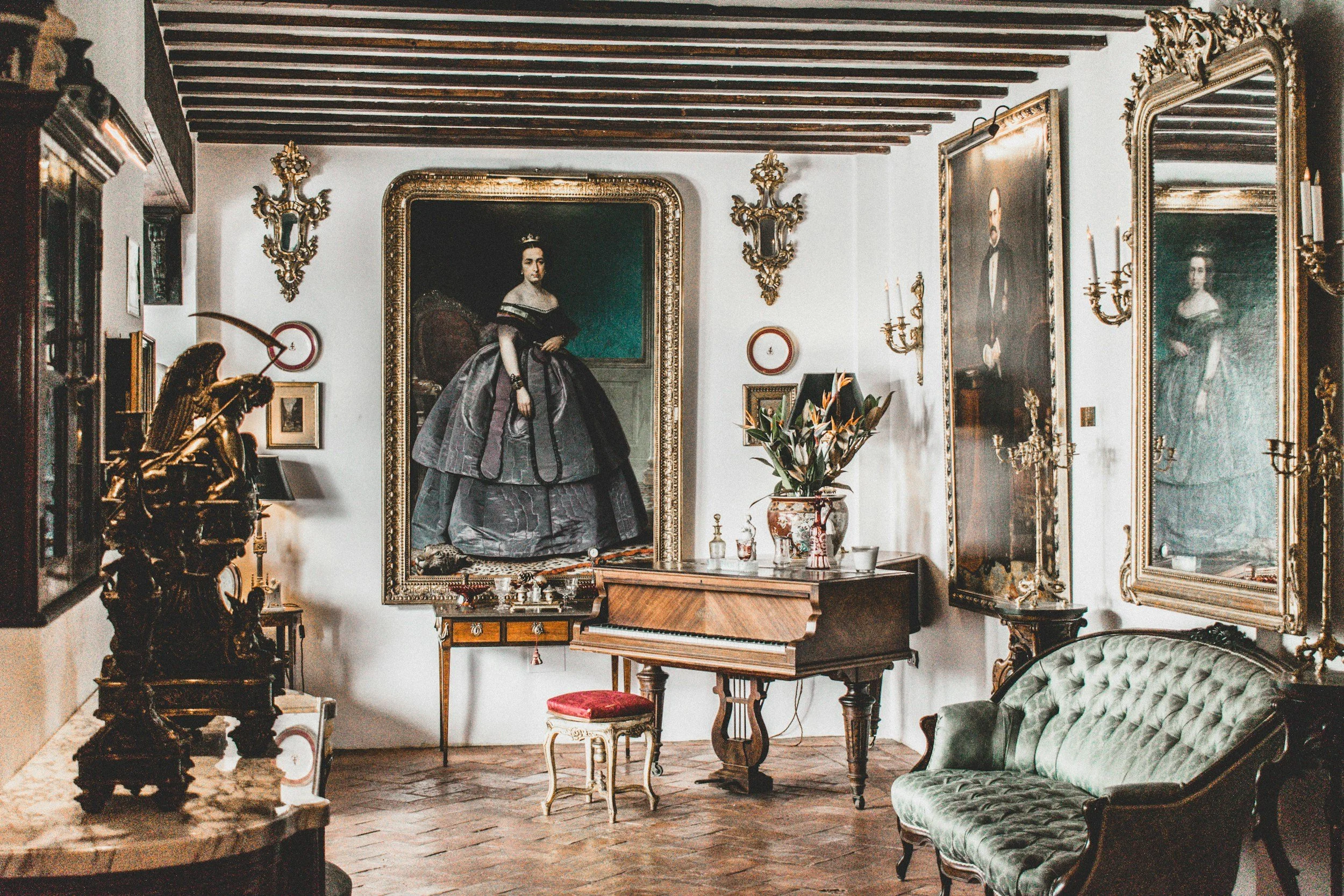 A vintage living room with ornate gold-framed portraits on the white walls, including a woman in a dark dress with a crown and a man in a tuxedo. The room features wooden ceiling beams, antique wall sconces, a small table with a vase of flowers, a green velvet sofa, a wooden writing desk, and decorative sculptures and artwork.