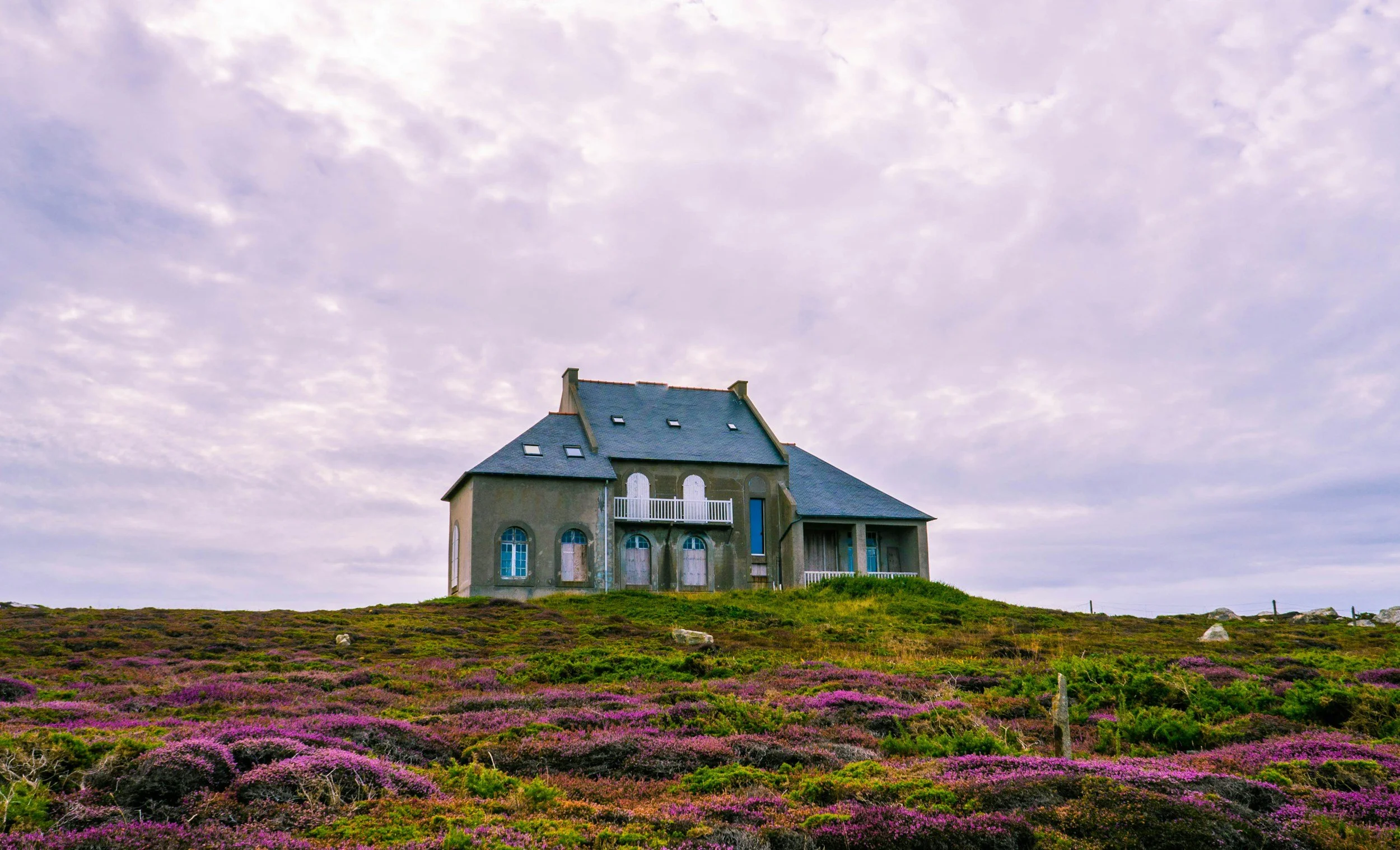 A house with a steep roof and multiple windows is situated on a grassy hill, with purple flowers and green bushes in the foreground, and a cloudy sky above.