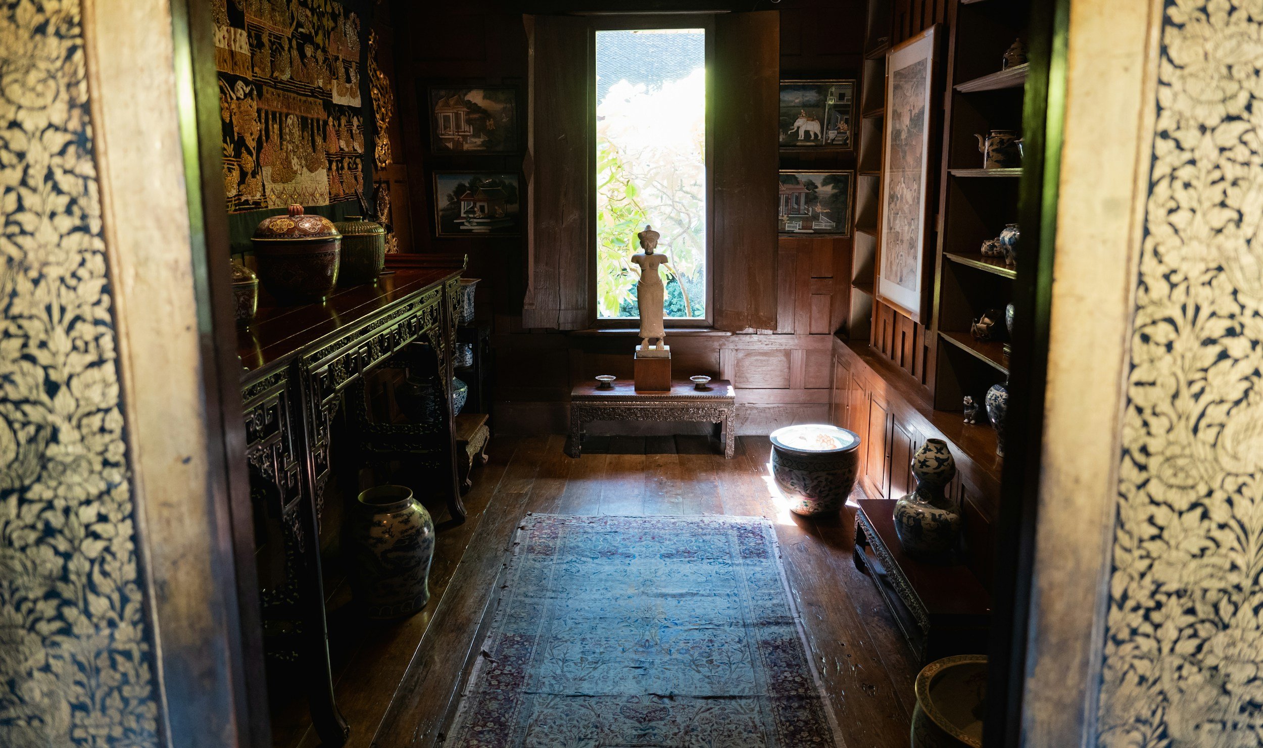 View of a cozy, vintage-style room with wooden floors and walls, decorated with artwork and antique furniture. A small table with a sculpture in front of a window that lets in natural light, surrounded by vases and other decorative objects.