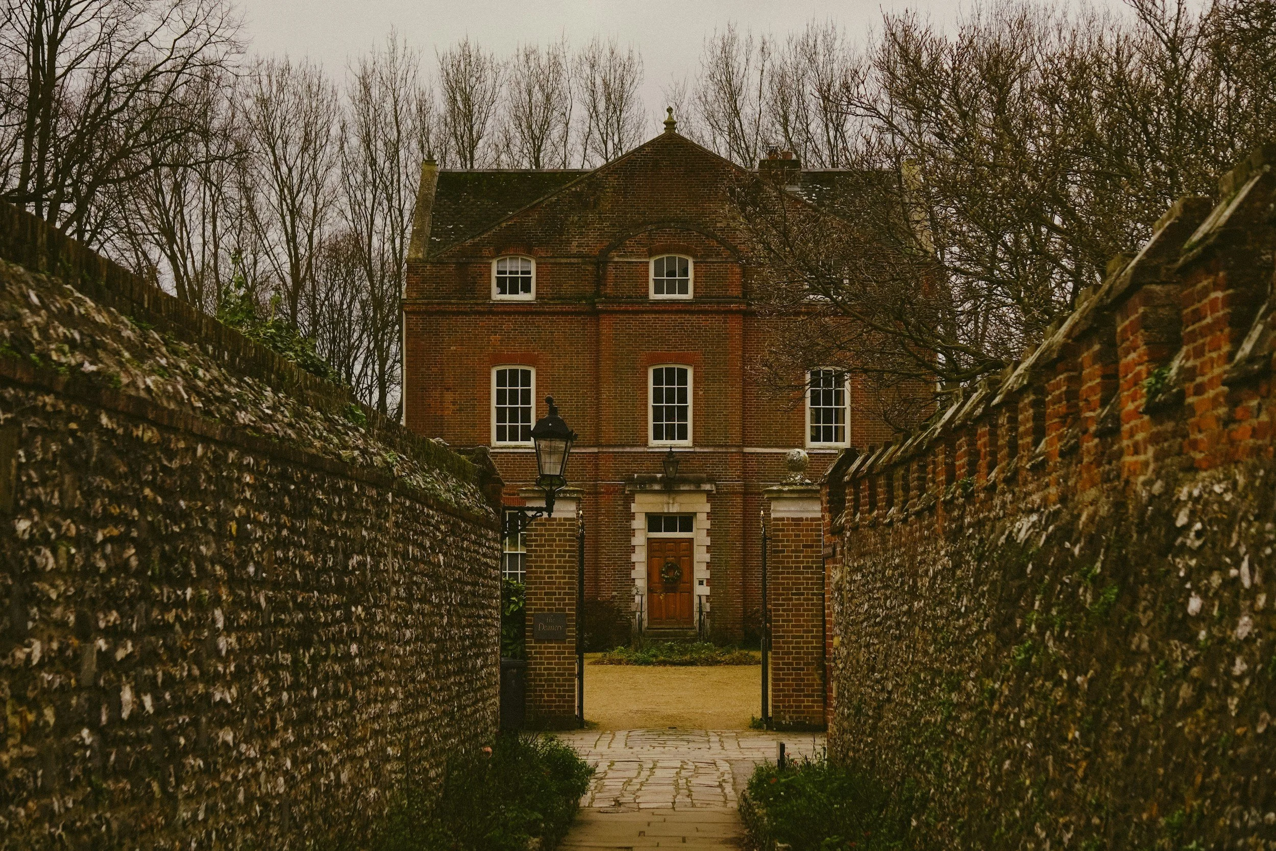 A brick house viewed through a stone-walled alleyway with leafless trees in the background and a lamppost in front.