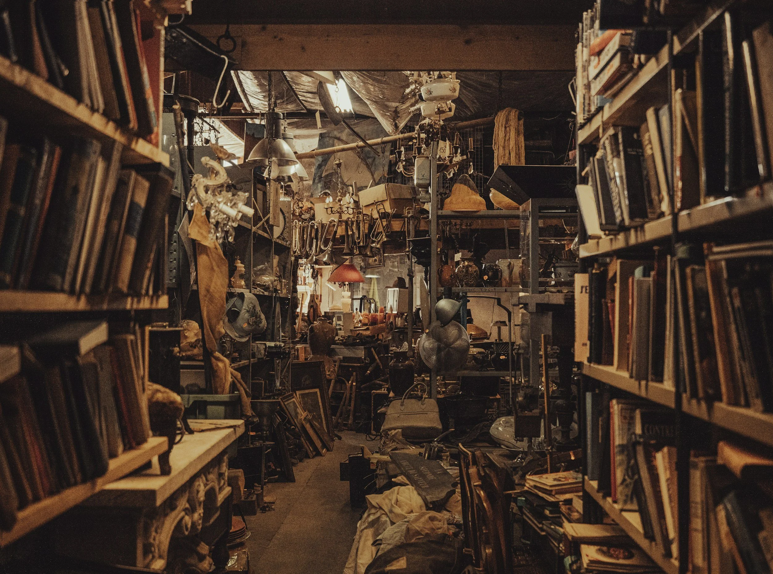 Interior of an old, cluttered bookstore or thrift store with narrow aisles, wooden shelves filled with books, and various scattered items and décor.