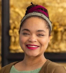 Close-up of a smiling woman with curly hair styled in a bun, wearing a headwrap and casual clothing, with a golden decorative background.