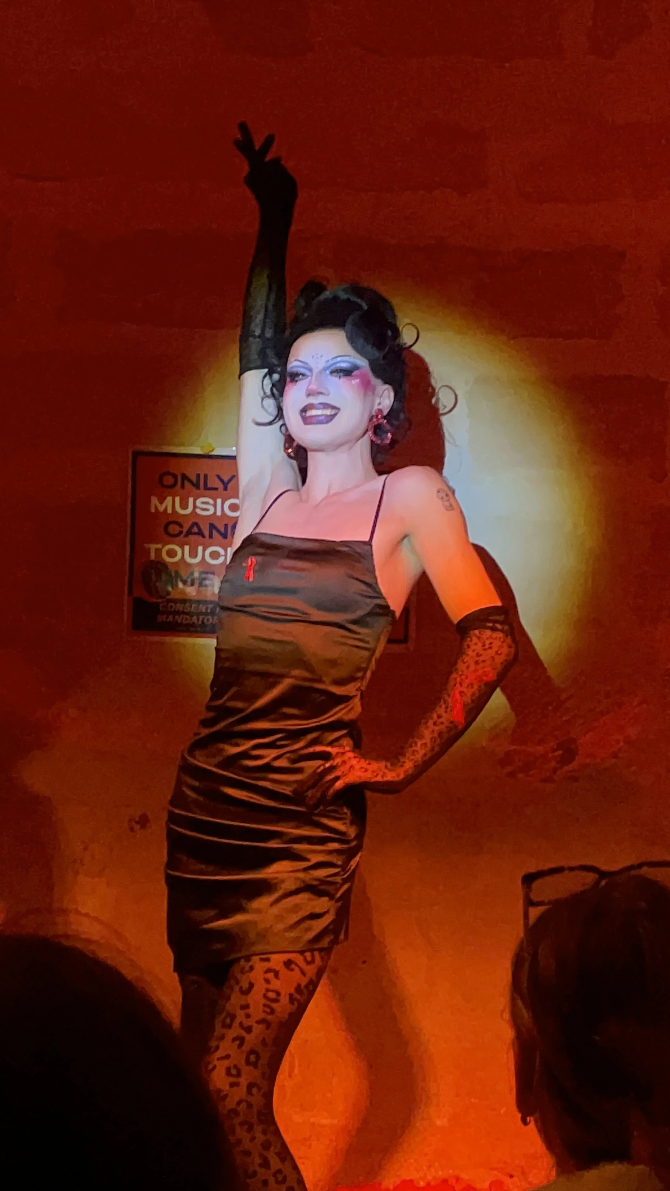 Une personne en costume de drag queen avec make-up coloré, robe en satin noir, et gants en dentelle à motifs léopard, posant sur scène avec un fond lumineux jaune-orange.