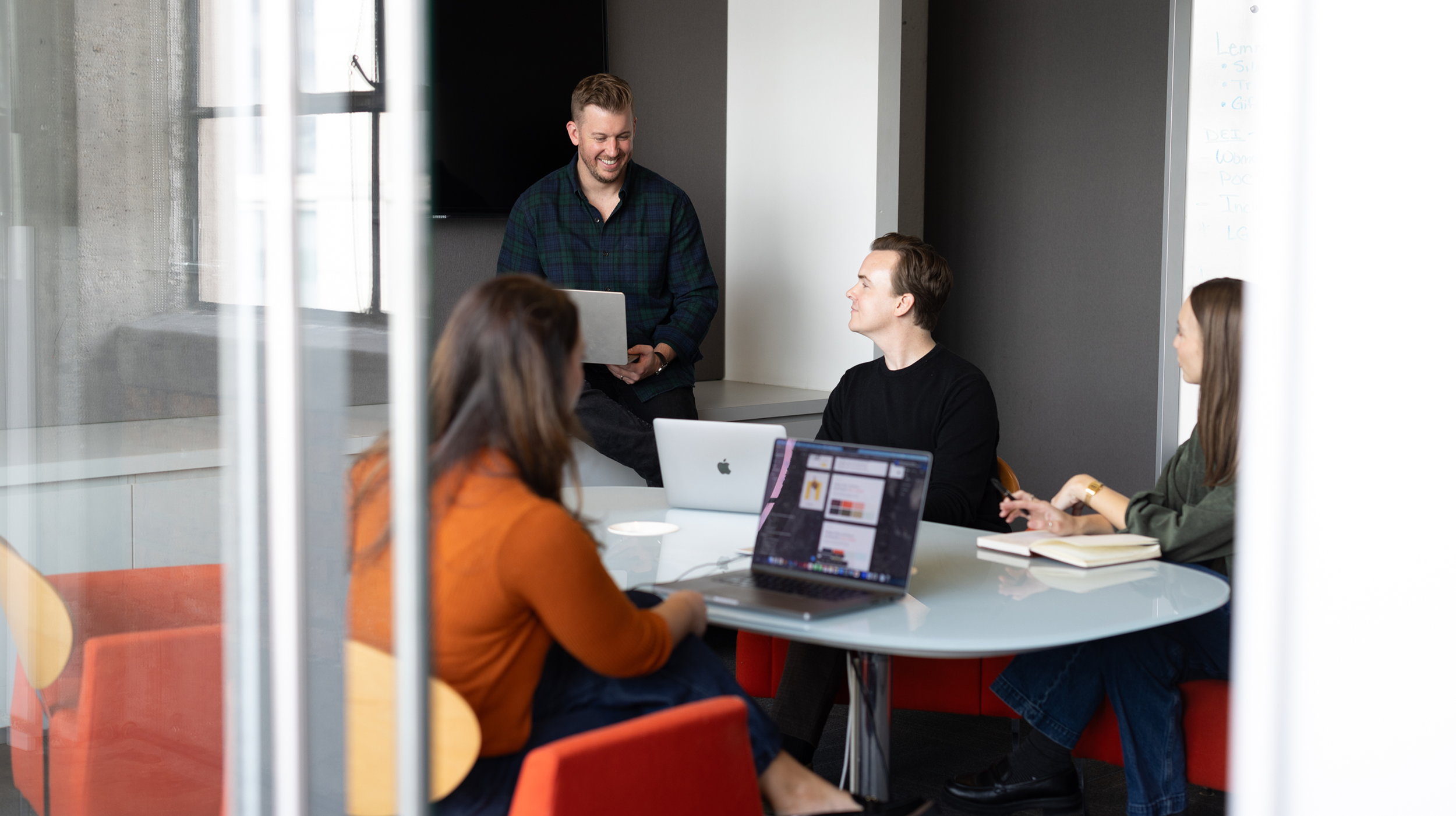 Group of Sunup employees in a modern office meeting room, engaged in a discussion. One man standing with a laptop and smiling, three others are using laptops and notebooks, with a whiteboard with notes in the background.